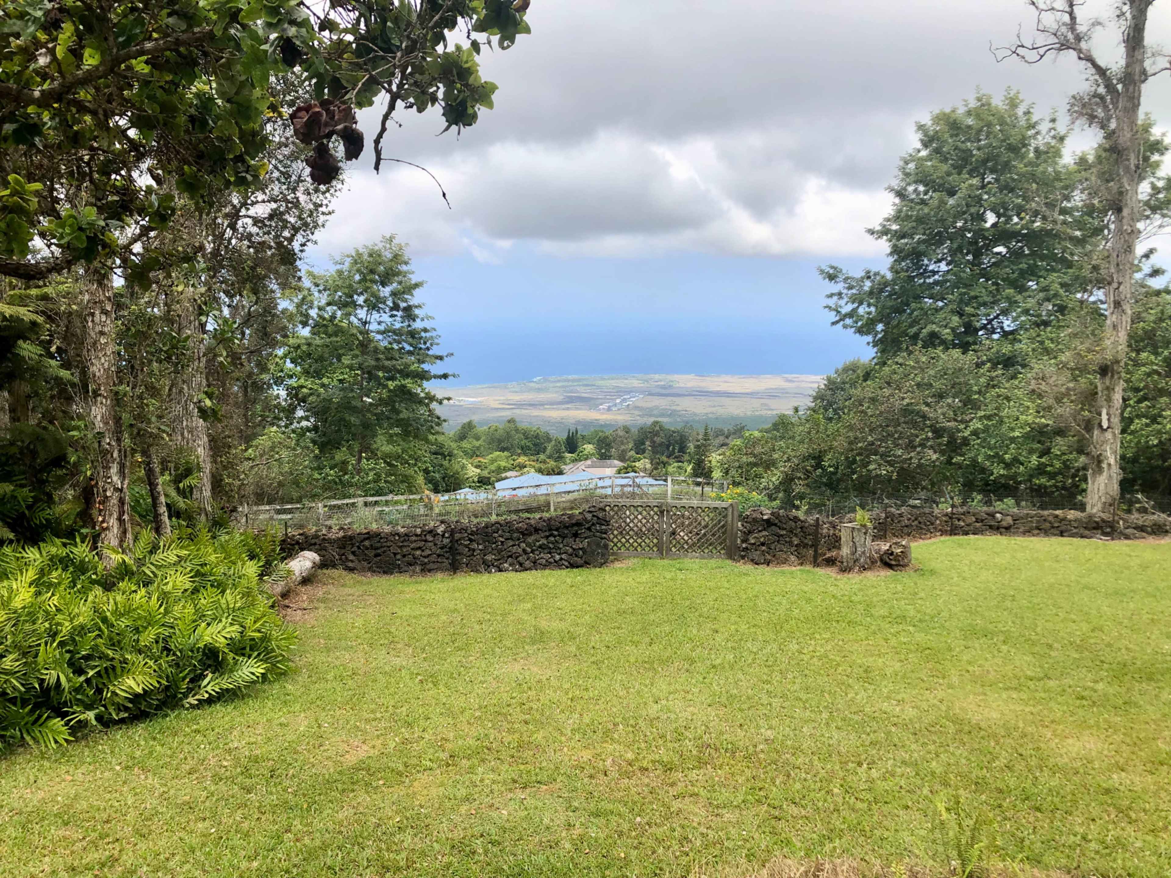 A grassy area bordered by trees, overlooking a landscape with distant hills under a cloudy sky.