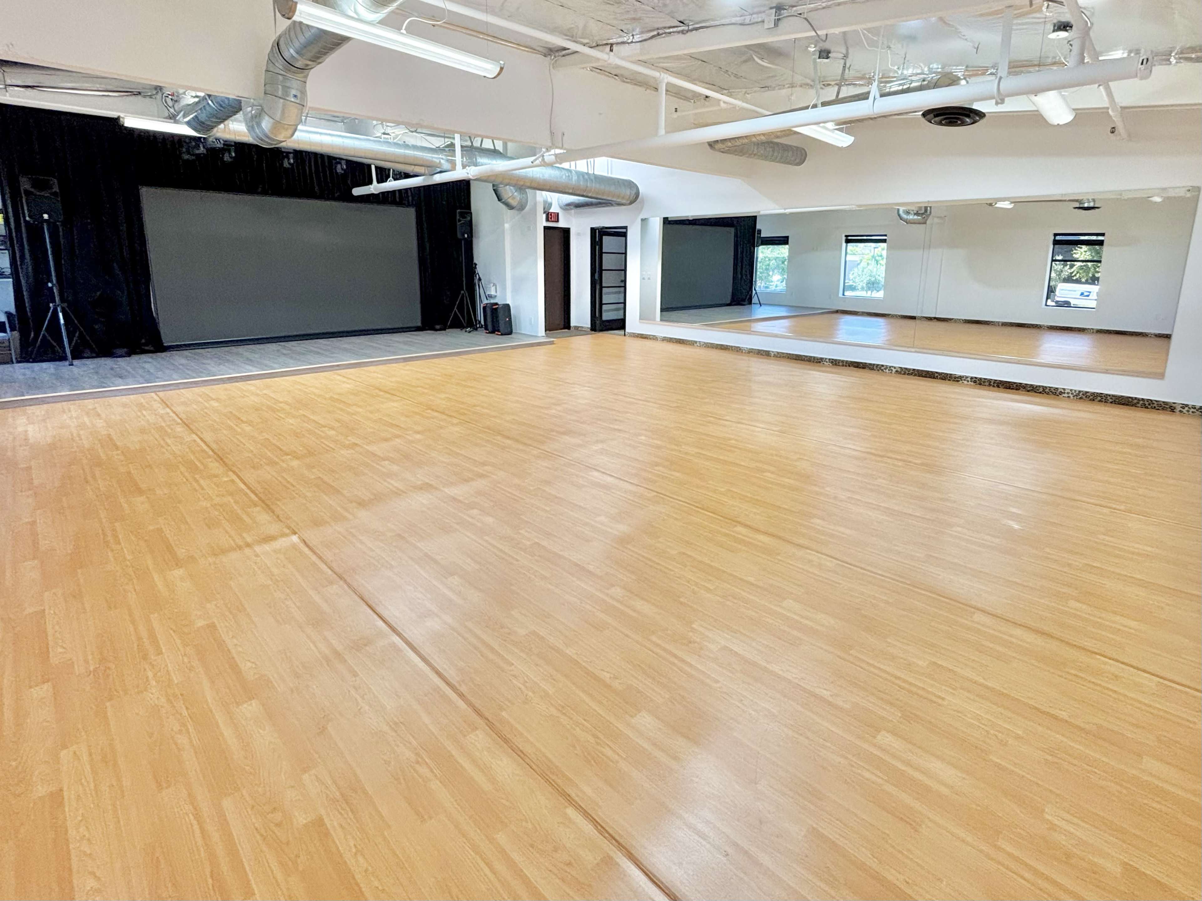 The image shows an empty studio space with a wooden floor, large mirrors on one side, and a black screen area at the front.