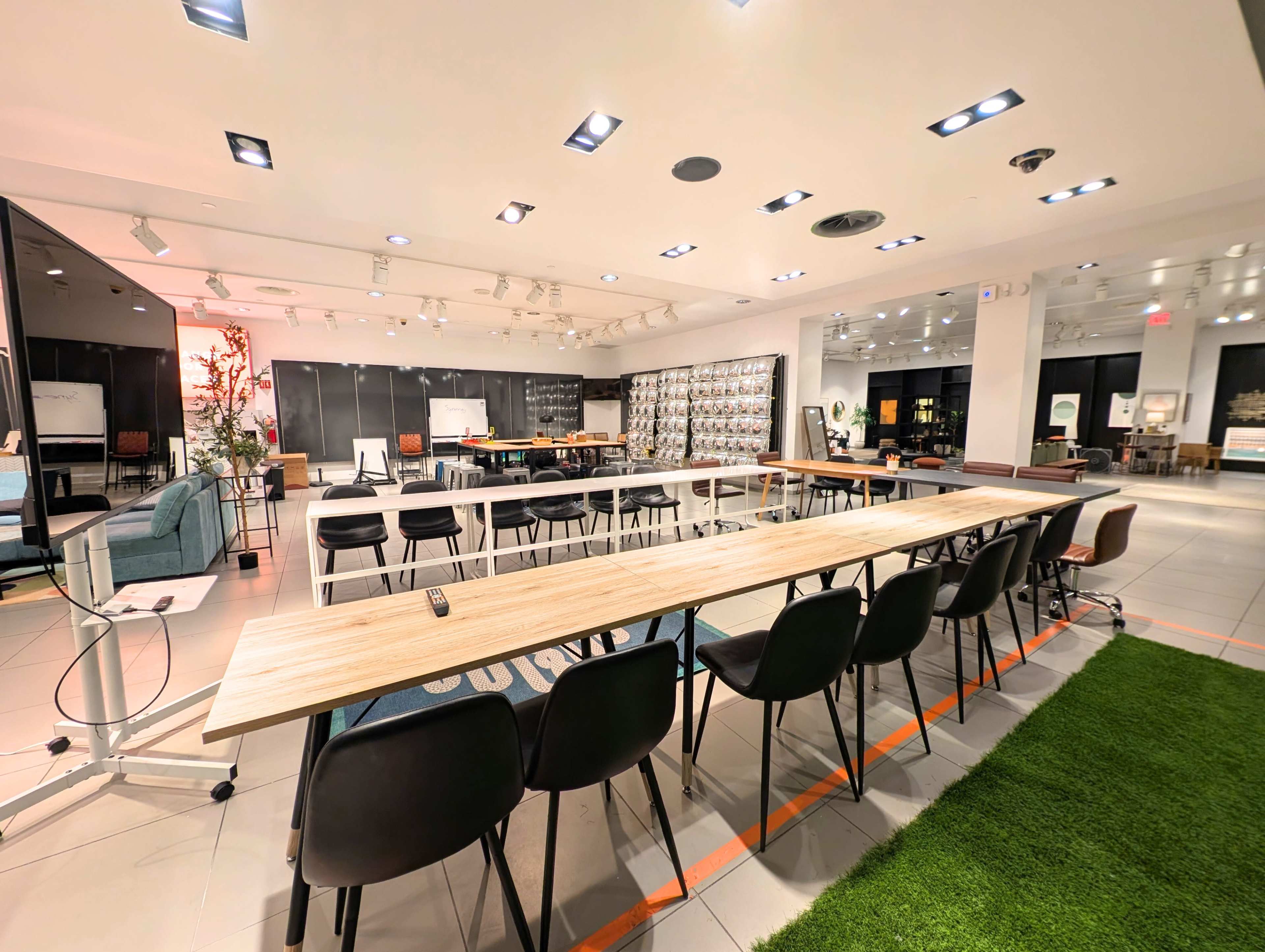 The image shows a modern conference room setup with a long wooden table surrounded by black chairs, featuring ample lighting and various furnishings arranged around the space.