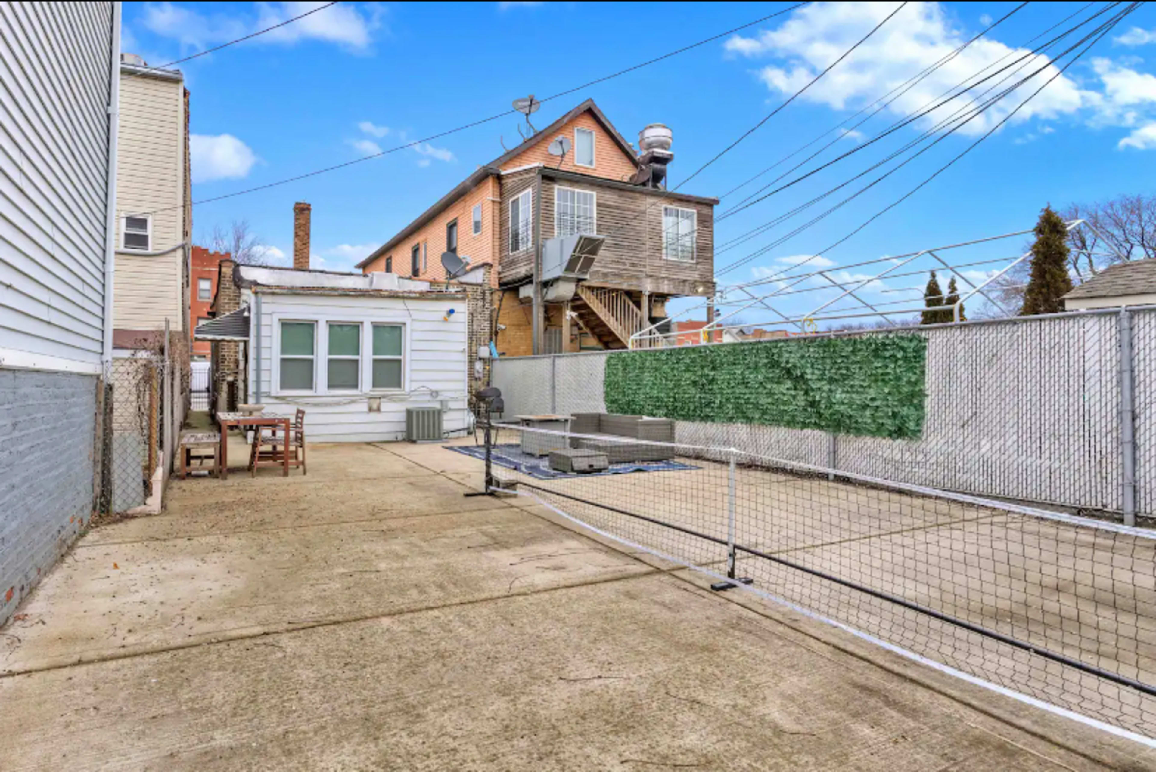The image shows a concrete backyard area enclosed by a fence, with a small seating arrangement and a building in the background.