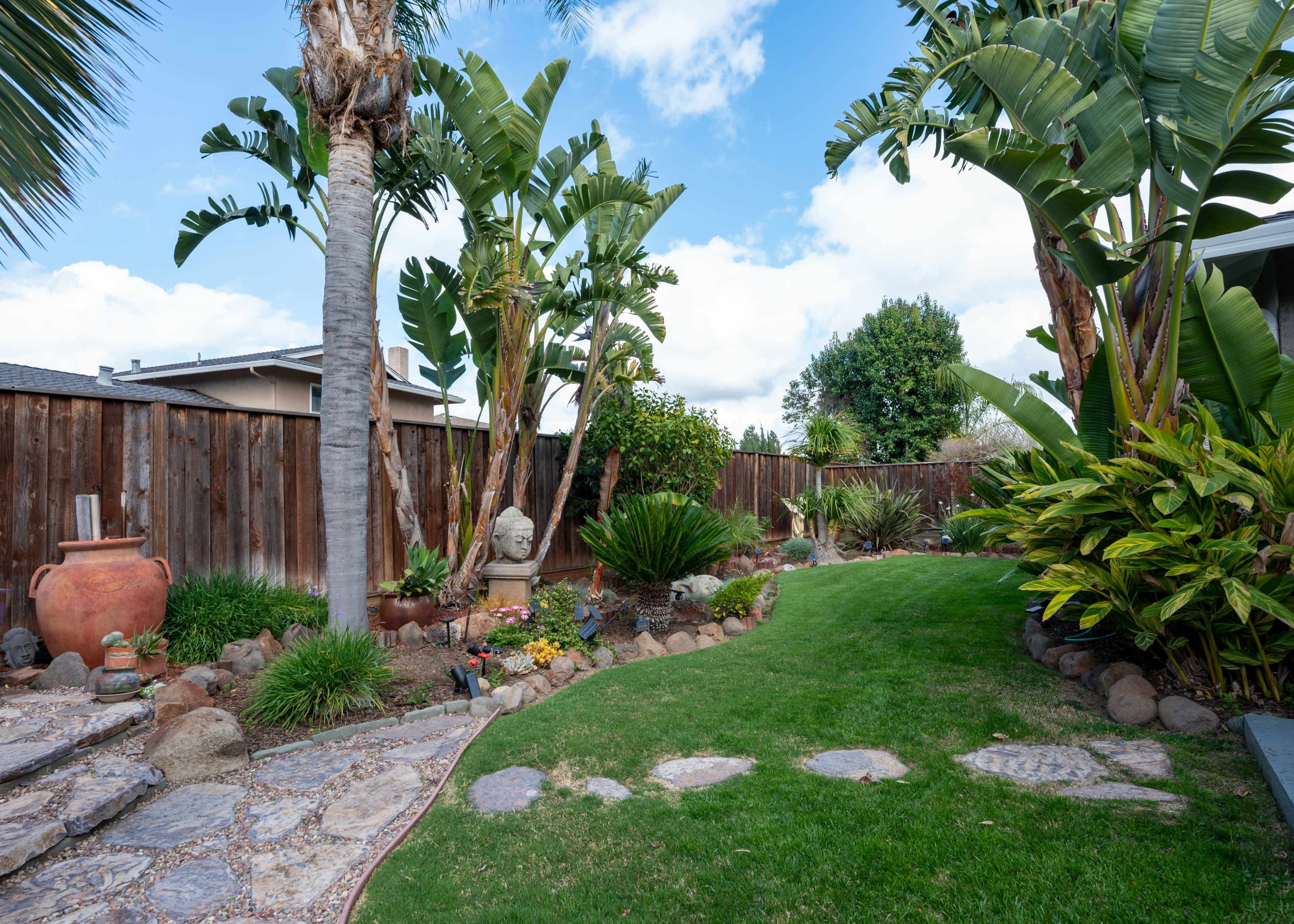The image shows a landscaped backyard with a winding stone path, tropical plants, and decorative elements, including a large terracotta pot and a stone statue.