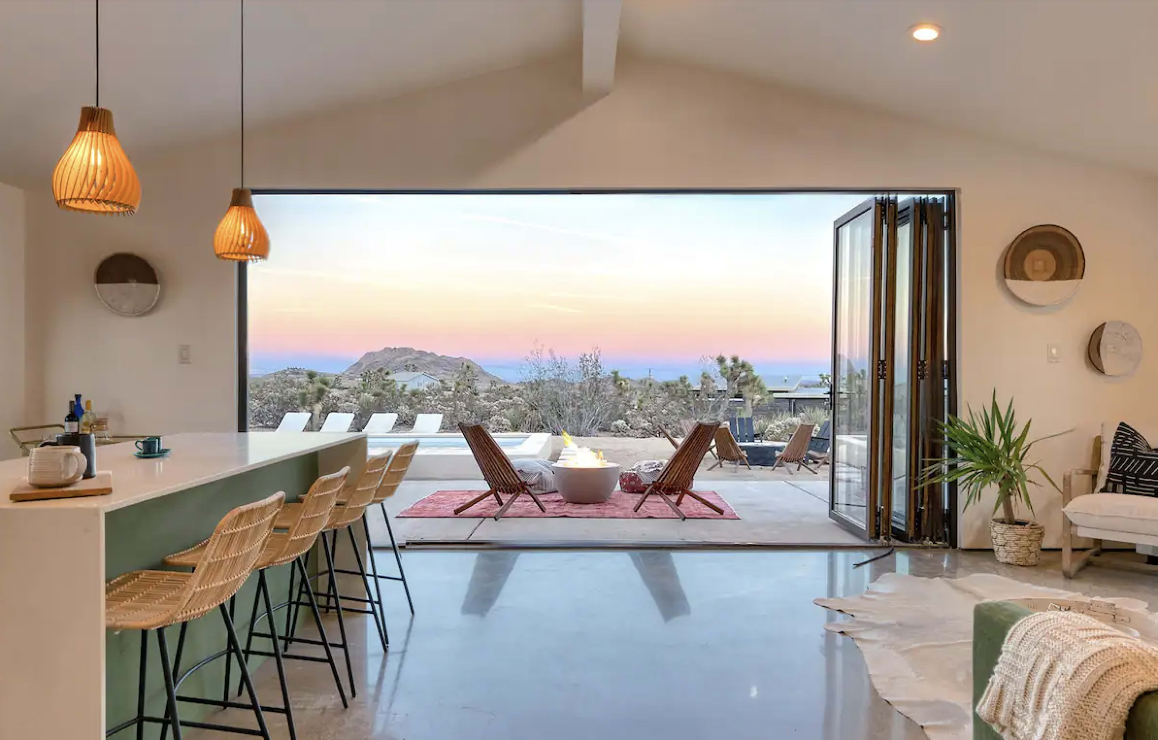 A modern interior kitchen opens to a patio with outdoor seating and a view of a mountain at sunset.