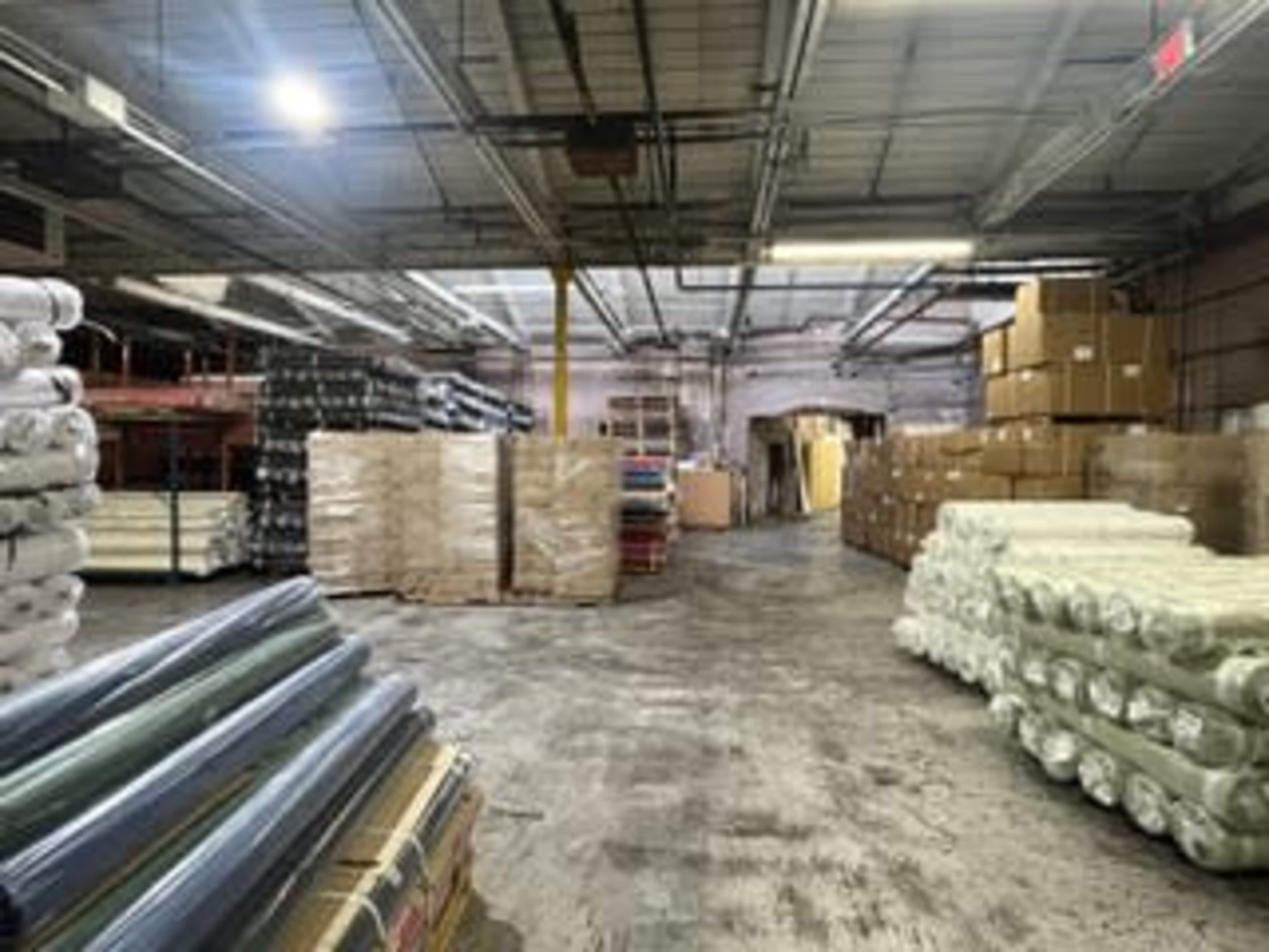 The image shows a large warehouse filled with rolls of material and stacks of cardboard boxes along the walls.