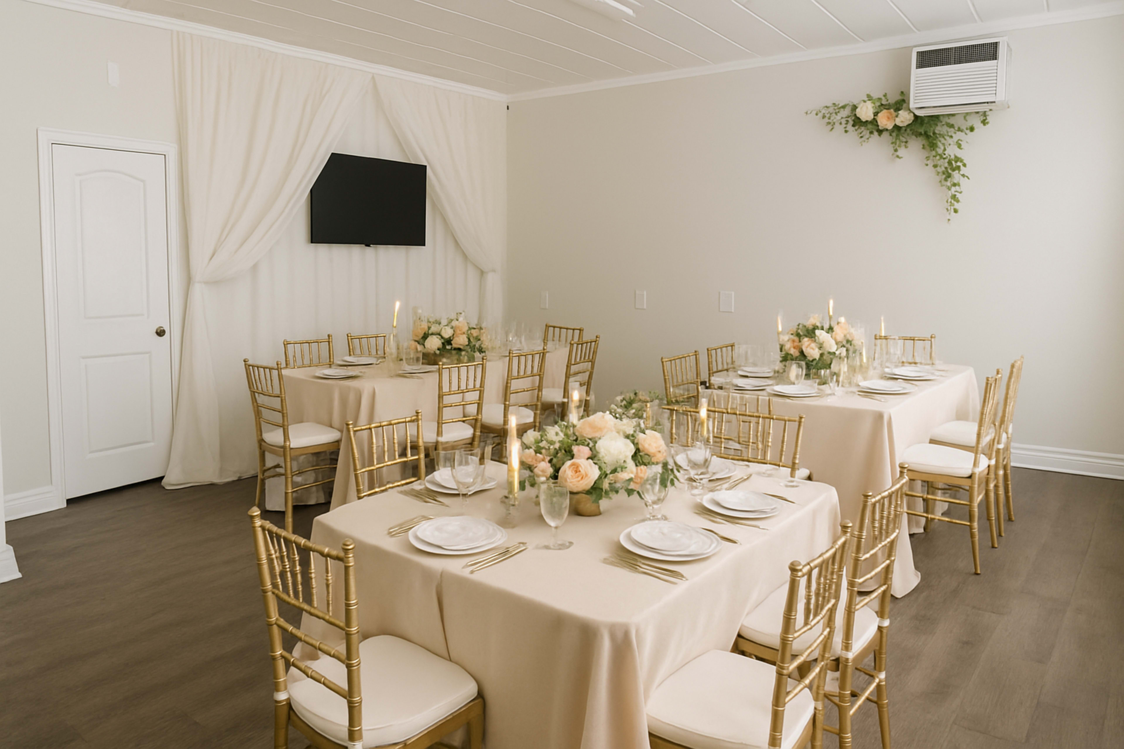 A well-decorated event space features two elegantly set dining tables with gold chairs and floral centerpieces under soft lighting.