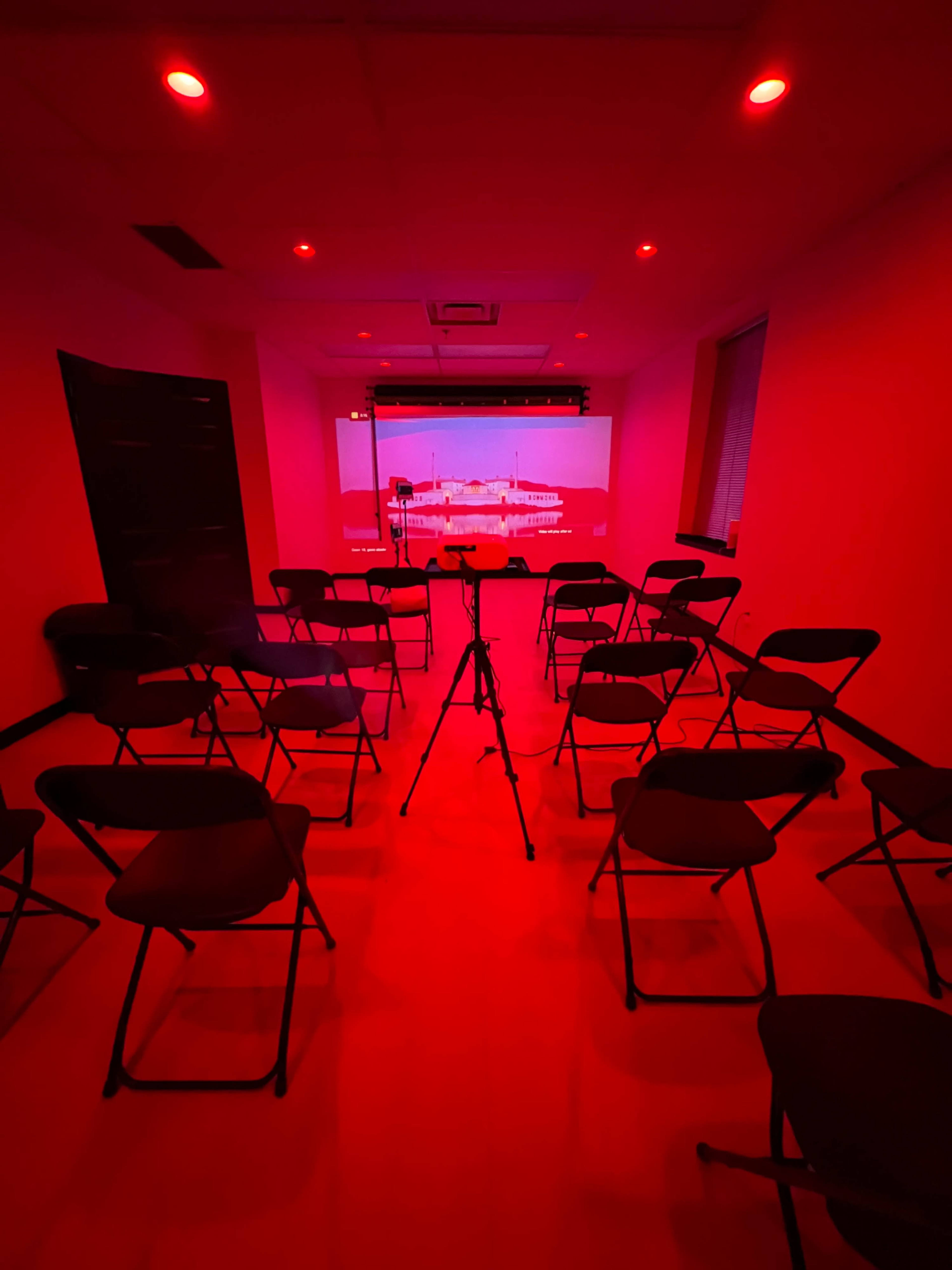 A presentation room with red lighting features arranged chairs in a circular formation around a projector screen displaying a red-tinted image.