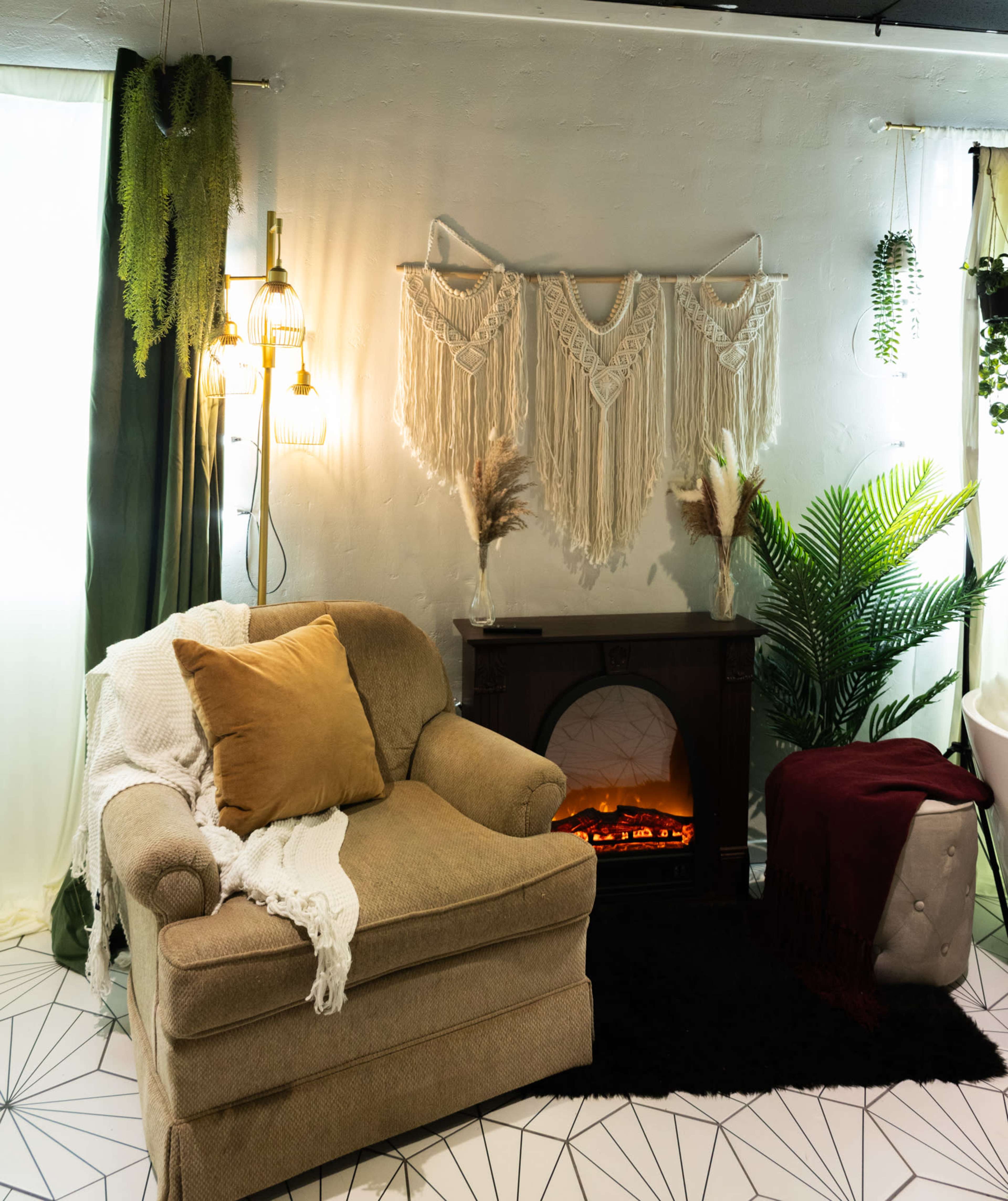 The image features a cozy living room corner with a beige armchair draped in a white throw, a faux fireplace with decorative elements, and various plants against a geometric-patterned floor.