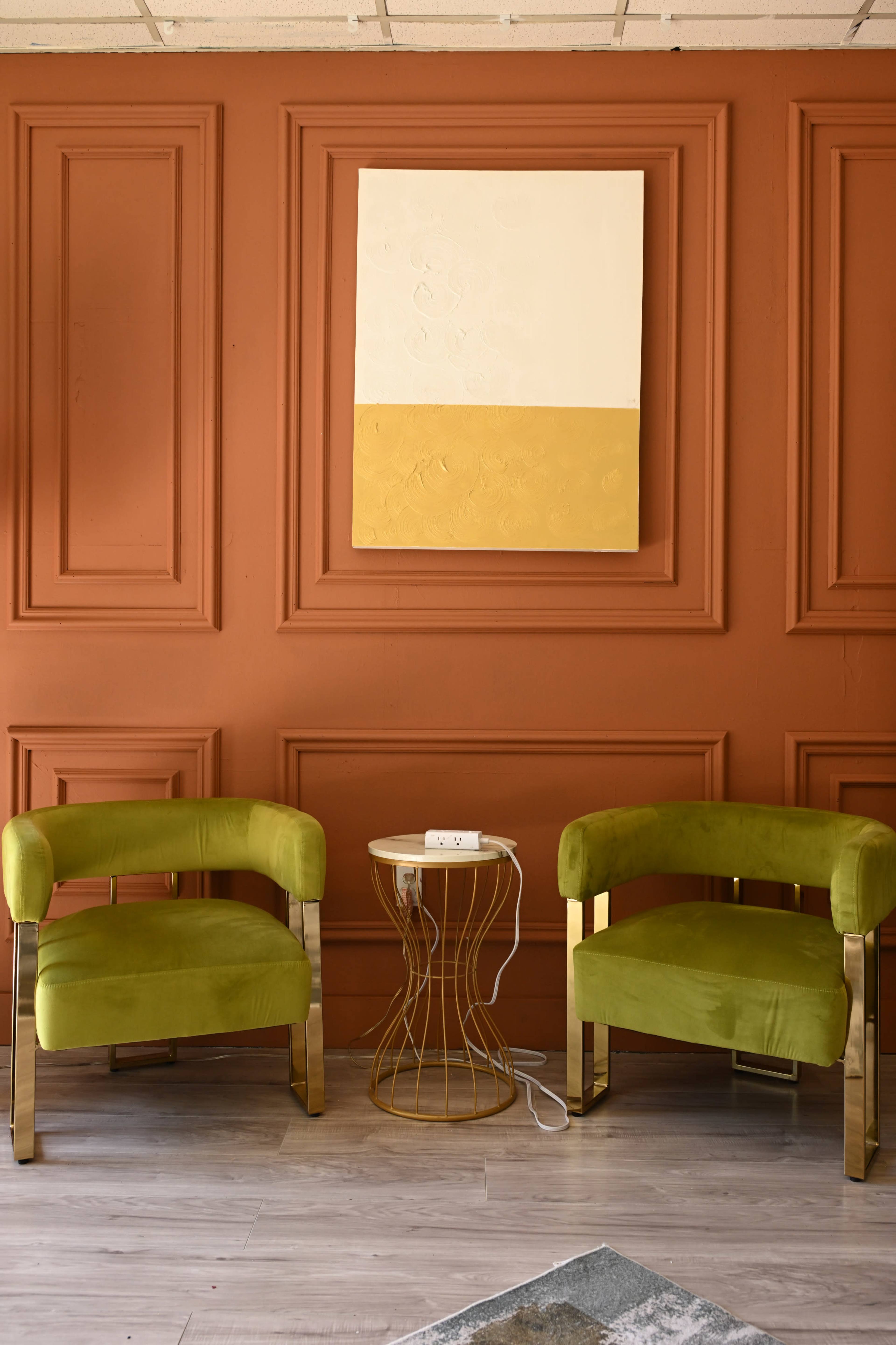 The image shows two green velvet chairs positioned on either side of a gold wire side table against a textured orange wall with a large abstract painting above.