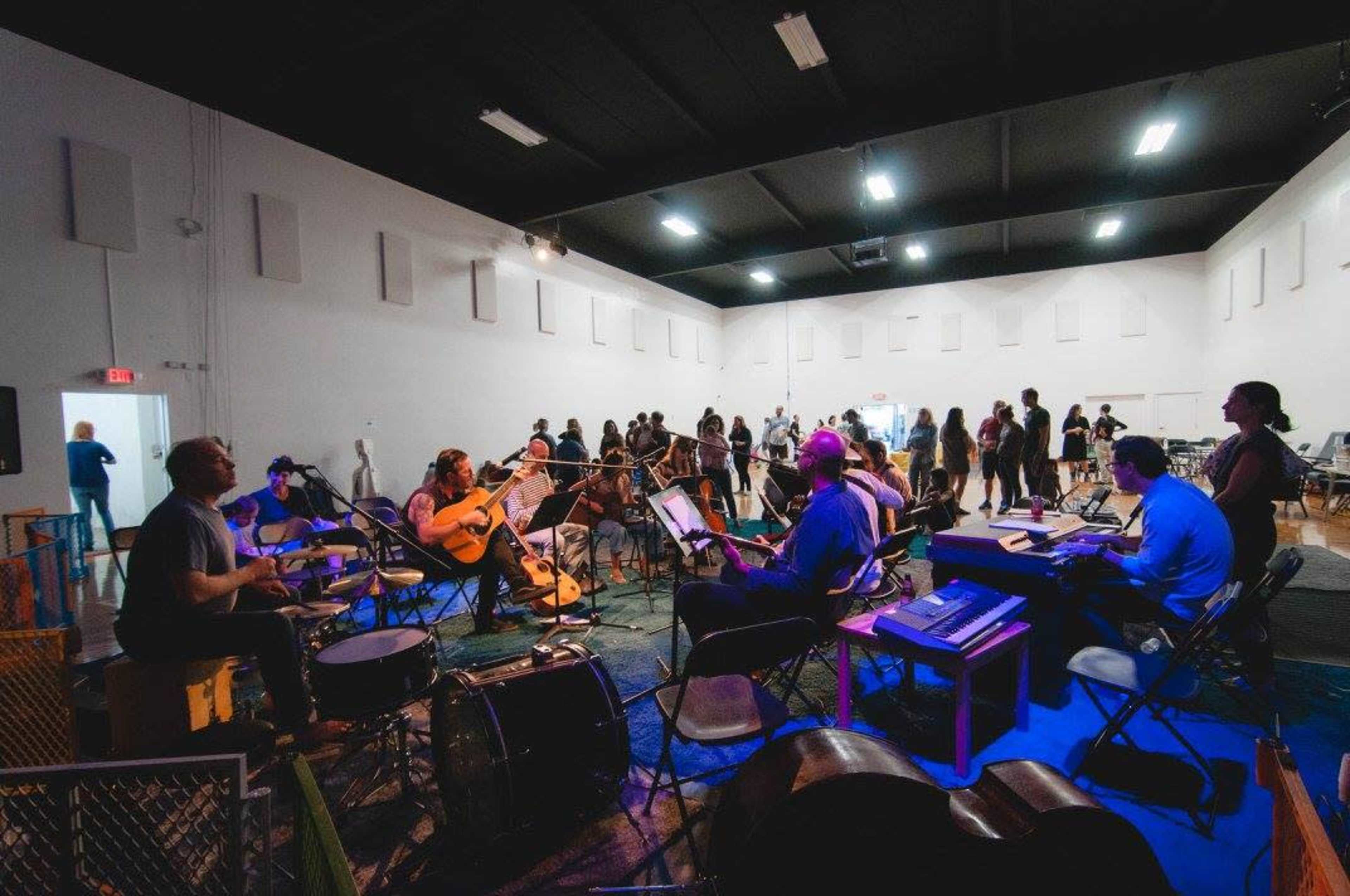 A group of musicians plays instruments in a spacious room while several people stand and mingle nearby.