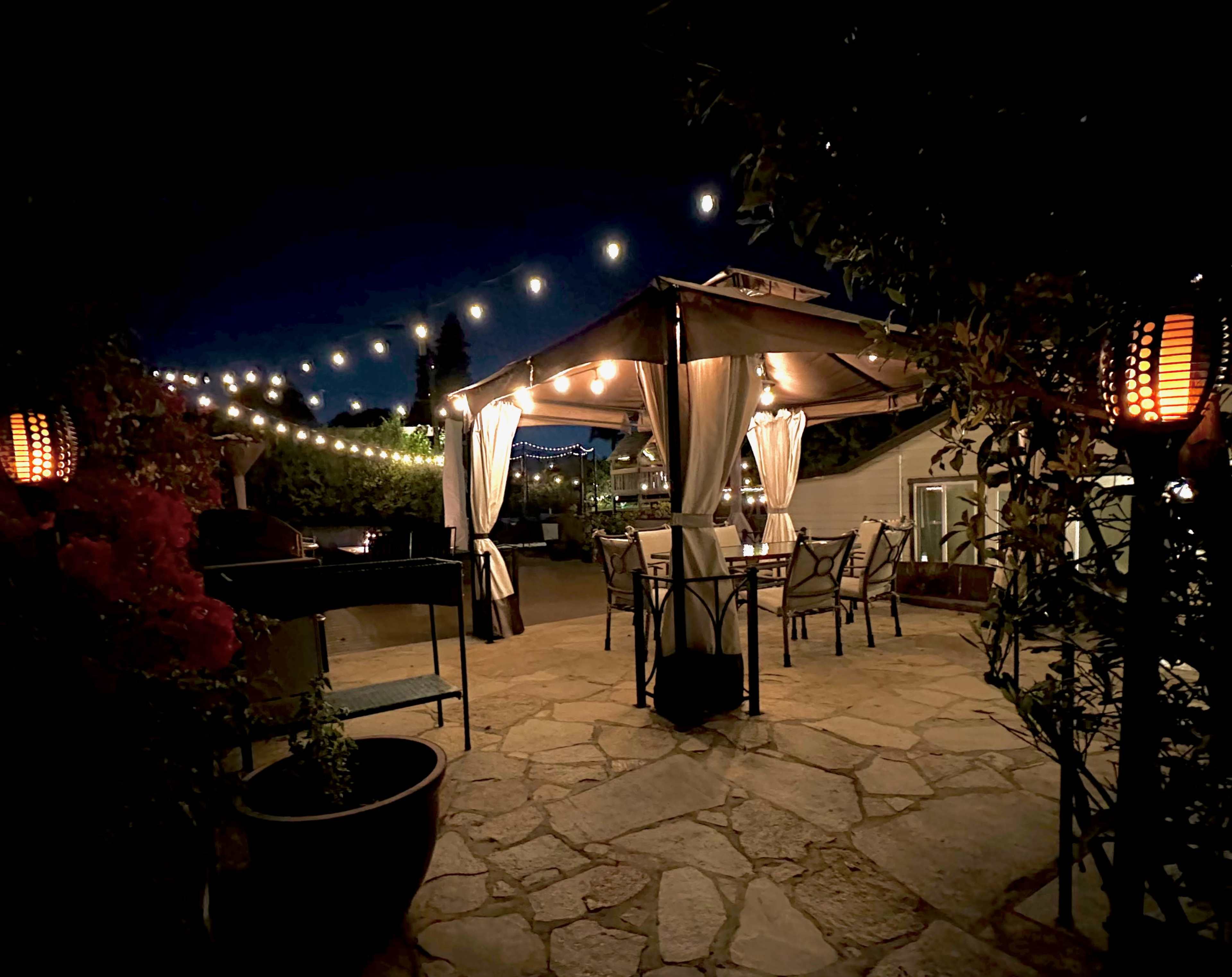 A stone-paved outdoor dining area is illuminated by string lights and lanterns, featuring a canopy over tables and chairs.