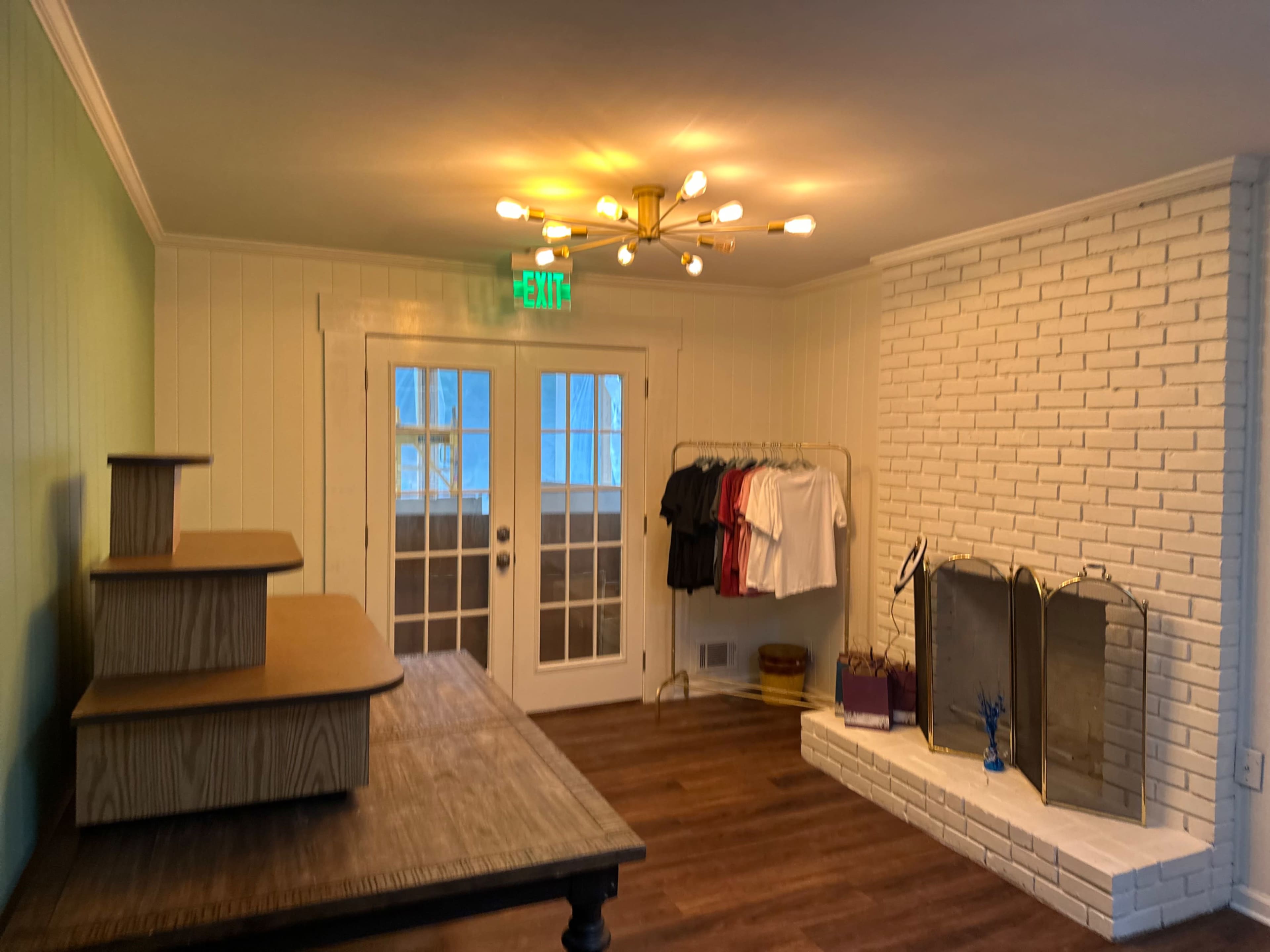 The room features a wooden table, a clothes rack with hanging shirts, a white brick fireplace, and large glass doors leading outside.