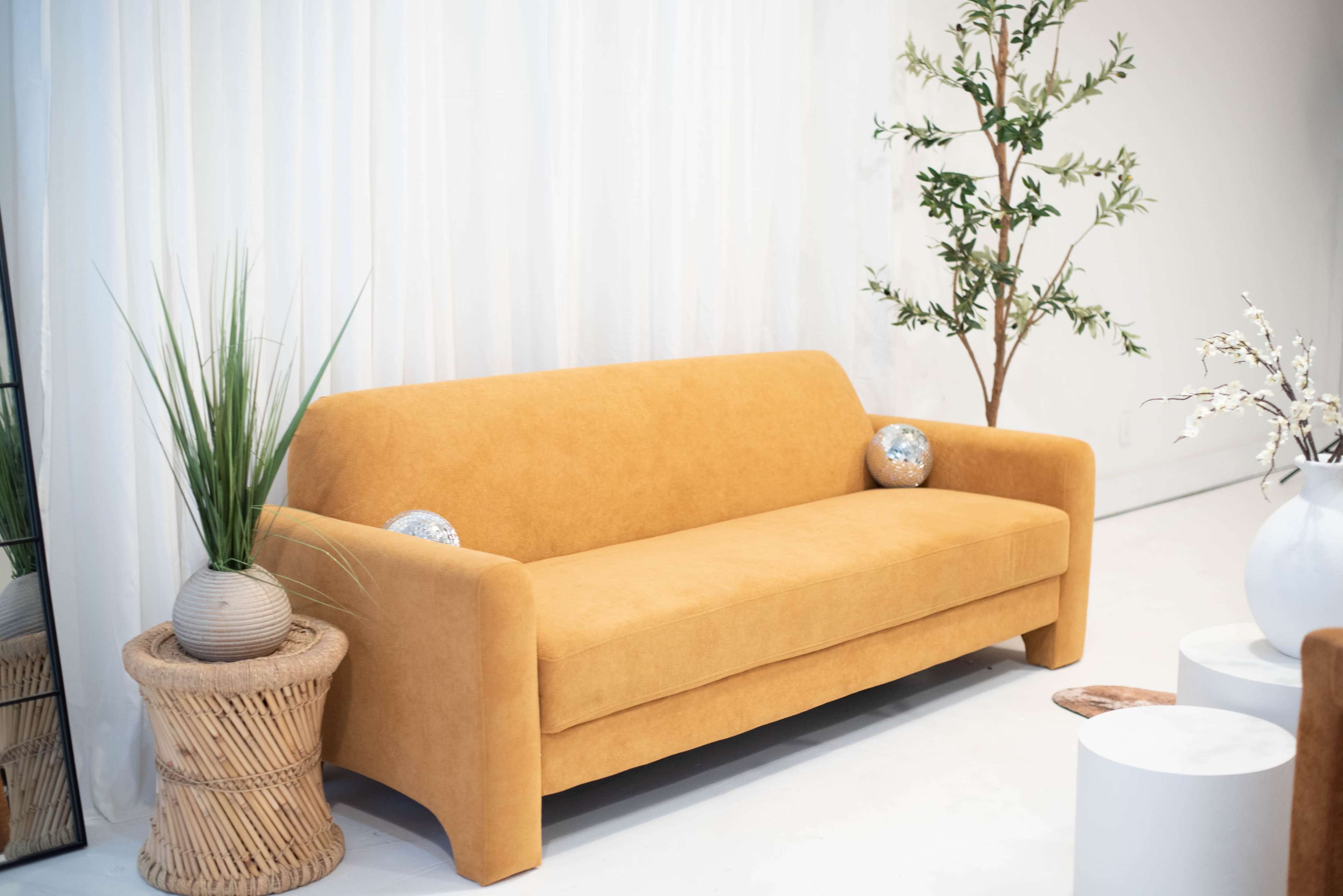 The image shows a mustard-colored sofa placed against a white curtain backdrop, accompanied by decorative plants and a mirror.