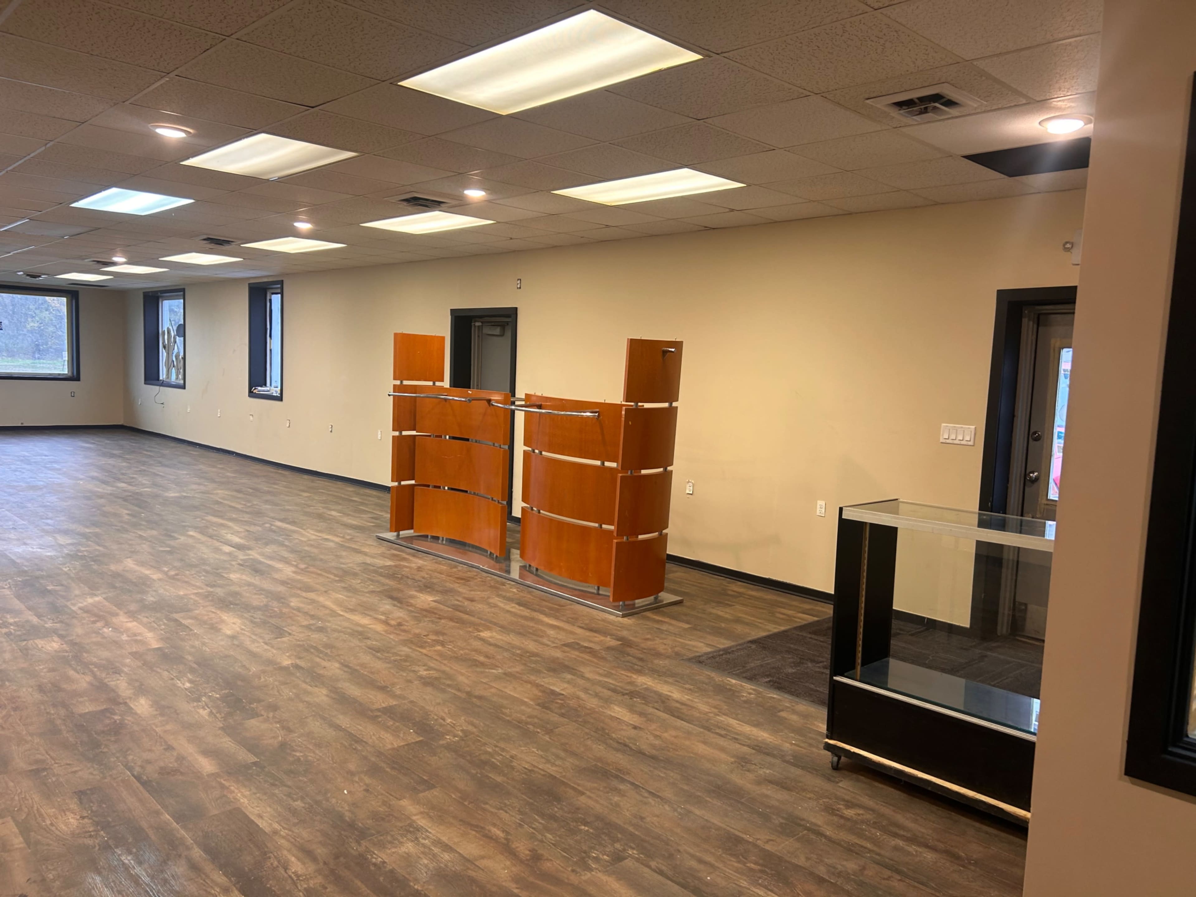 The image shows a spacious, empty room with laminate flooring, a reception area partition, and a glass display case.