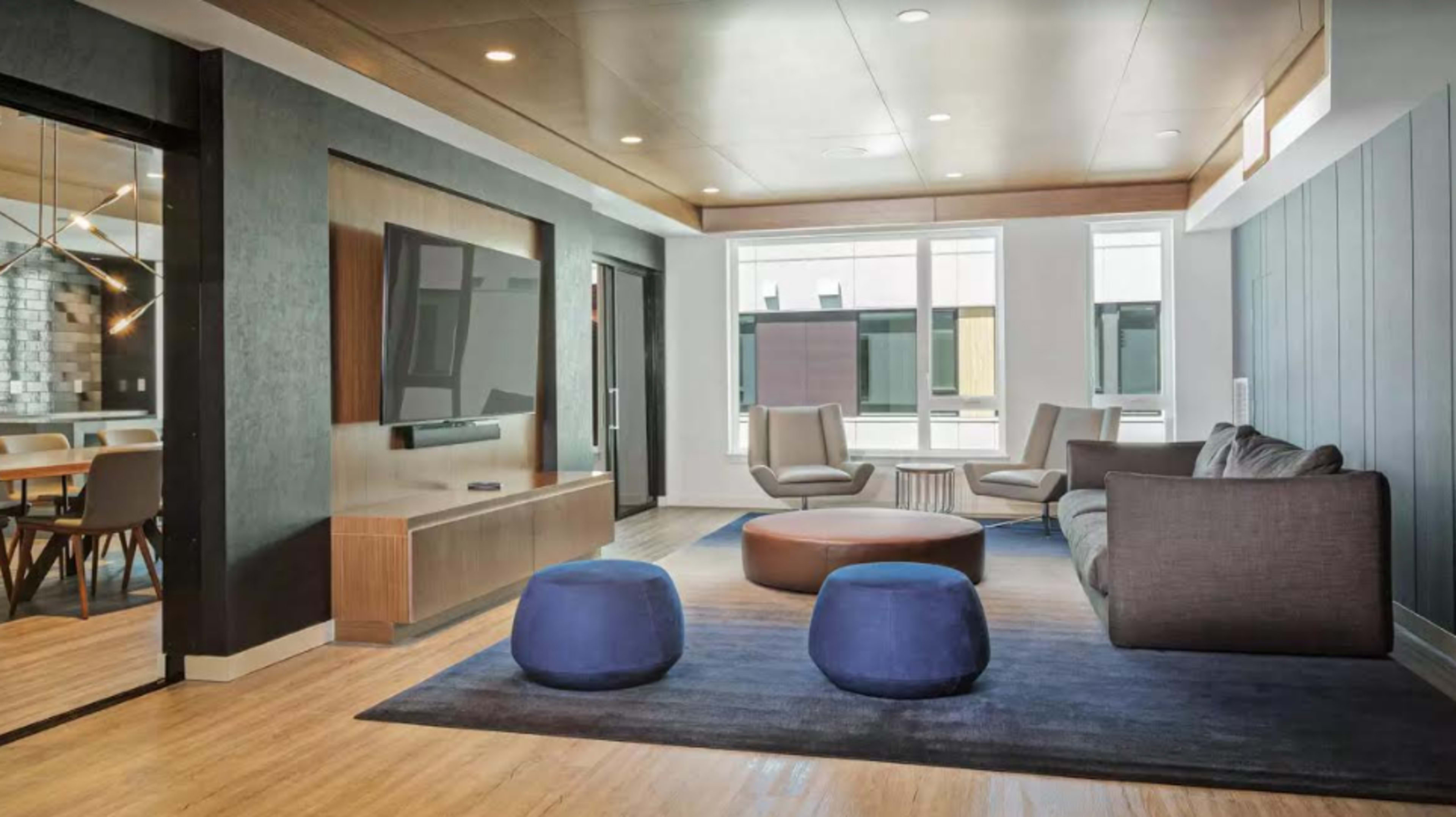 A modern lounge area with a gray sofa, two blue poufs, a wooden coffee table, and a television mounted on the wall, surrounded by large windows.