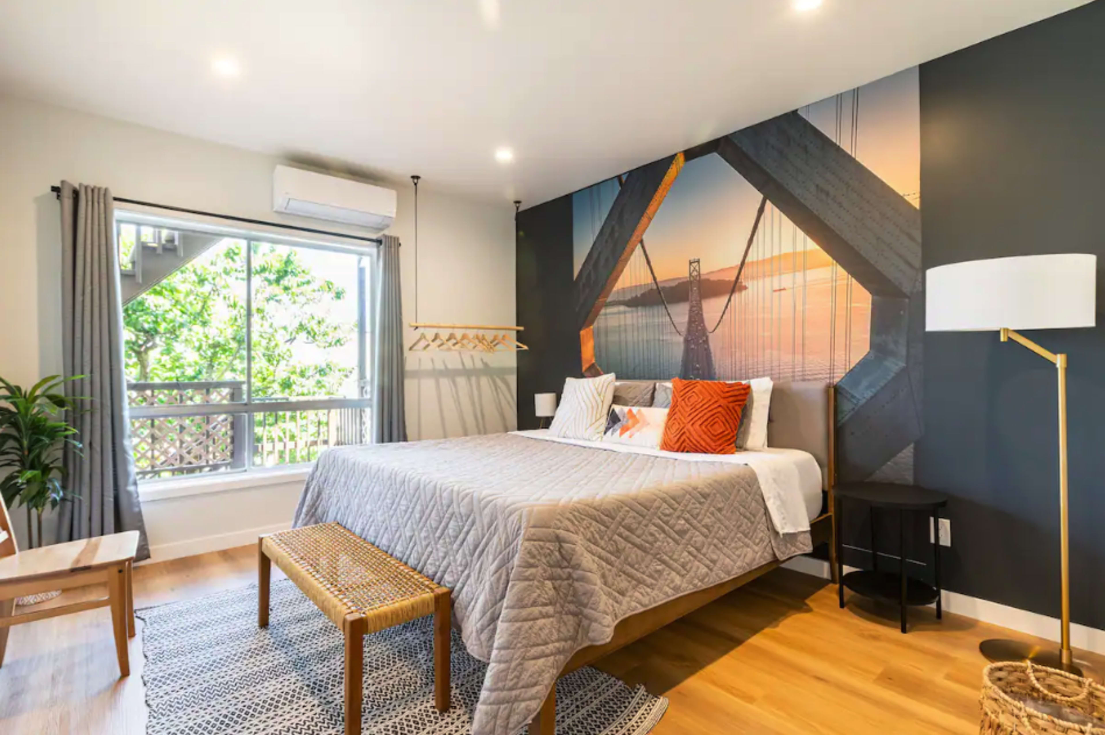 A modern bedroom features a large bed with a quilted blanket, complemented by a wall mural of a bridge and large windows allowing natural light.