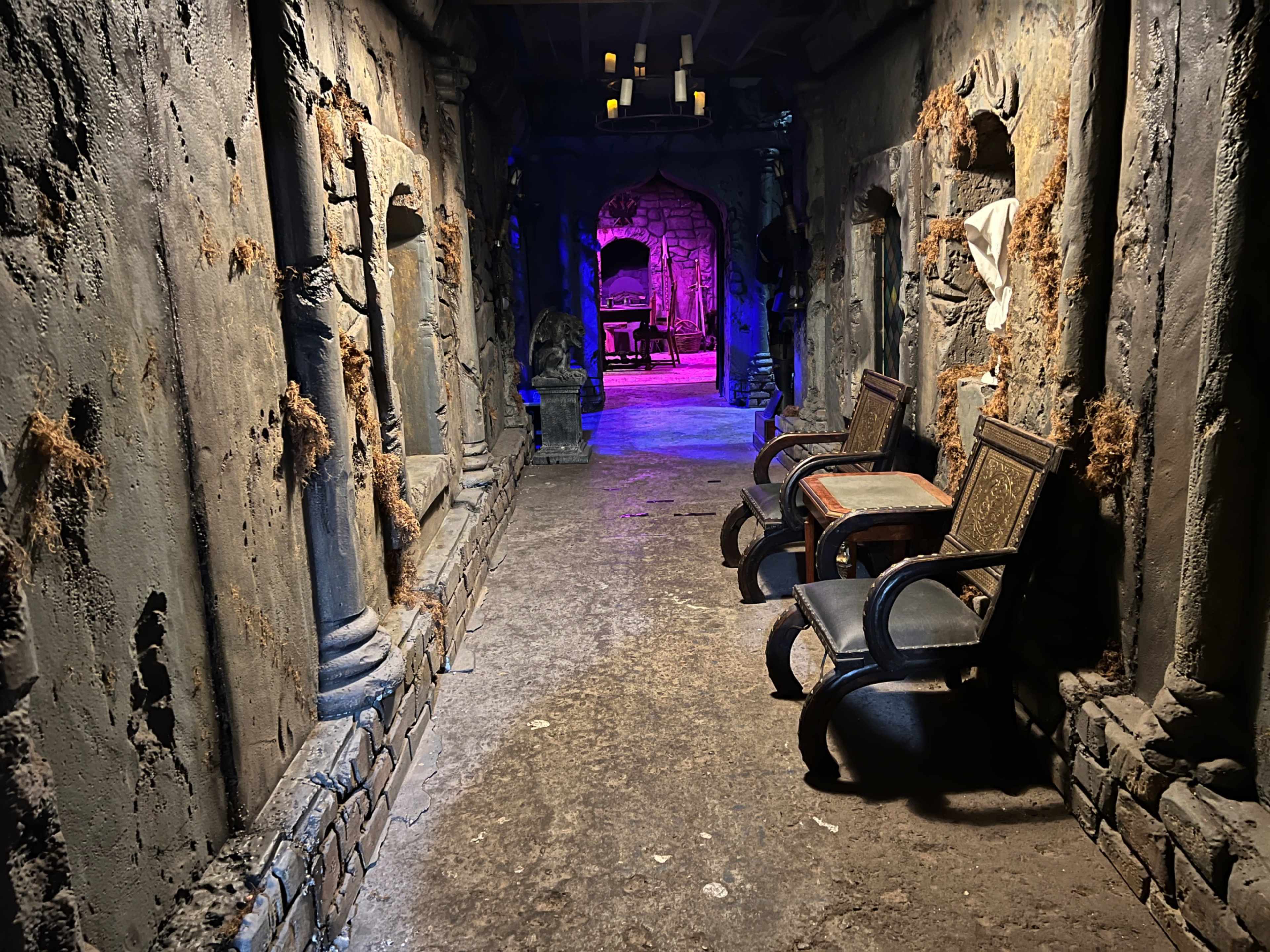 The image shows a dimly lit corridor with textured stone walls, featuring two wooden chairs and an entrance leading to a room illuminated in purple light.