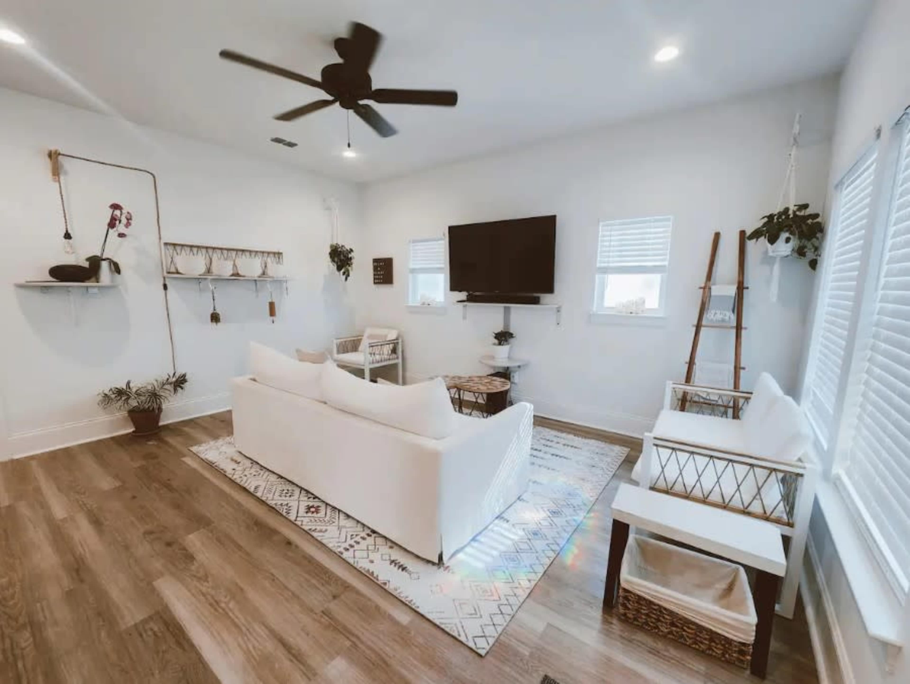 The living room features a white sofa, two chairs, a television mounted on the wall, and decorative plants arranged on shelves.