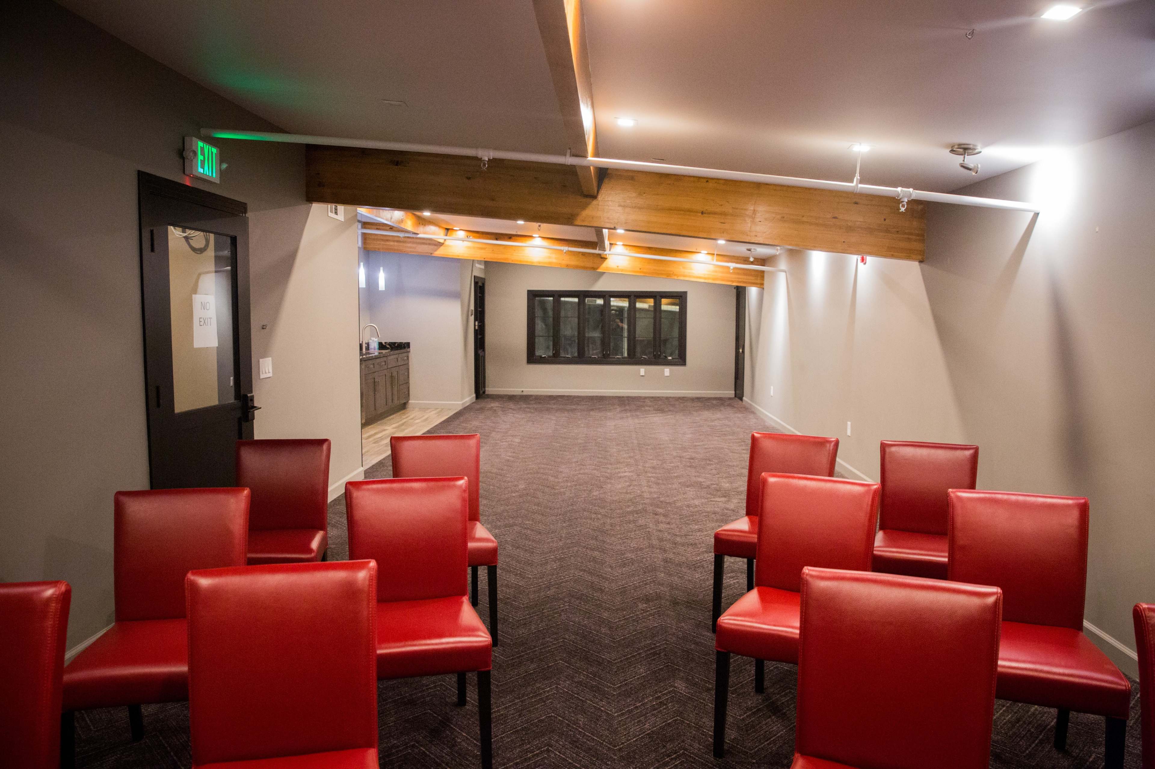 A conference room with red chairs arranged in rows, a wooden beam overhead, and a bar area in the background.