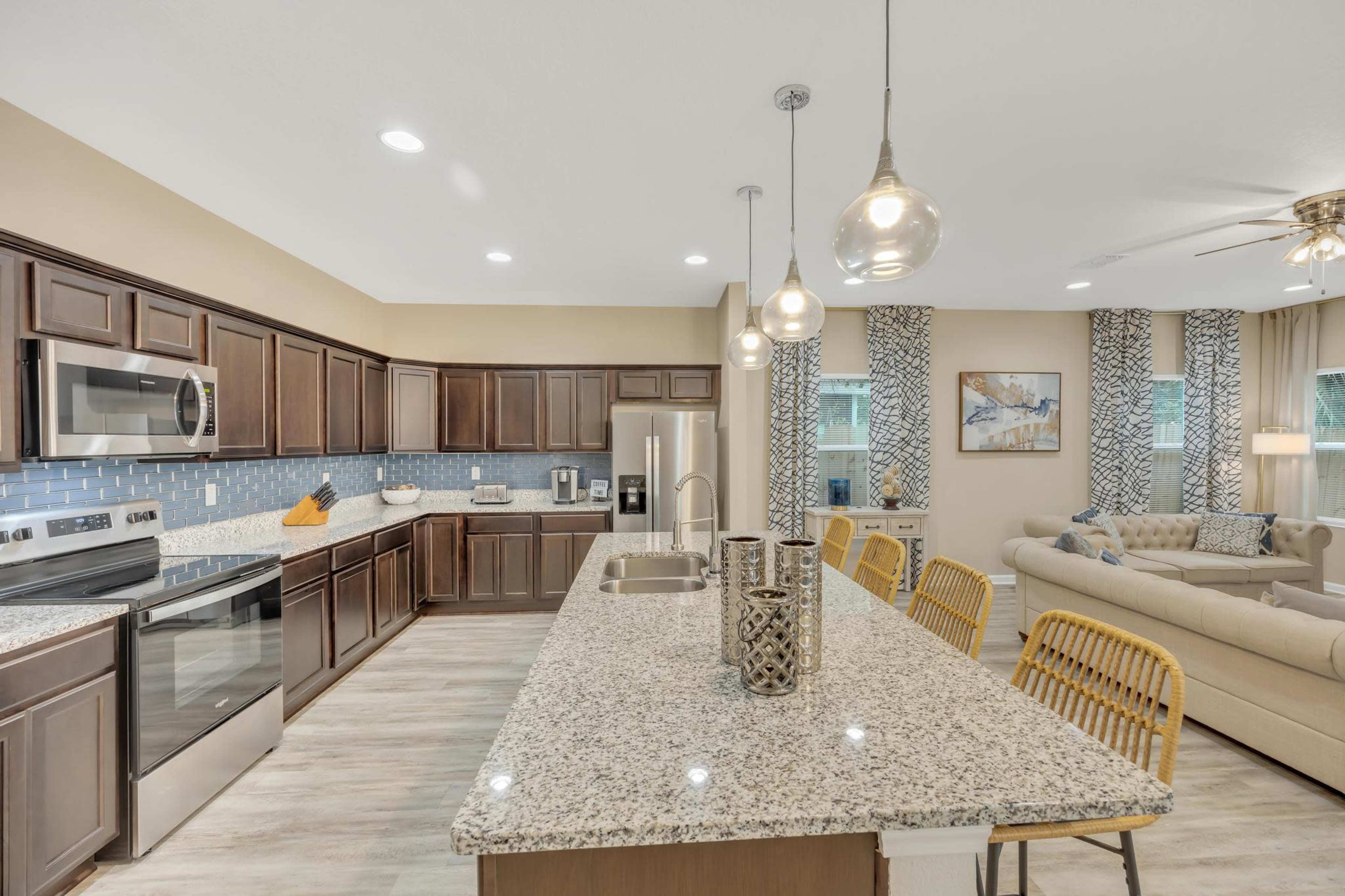 The image shows a modern kitchen with dark wooden cabinets, stainless steel appliances, and a spacious island with granite countertops, adjacent to a cozy living area.