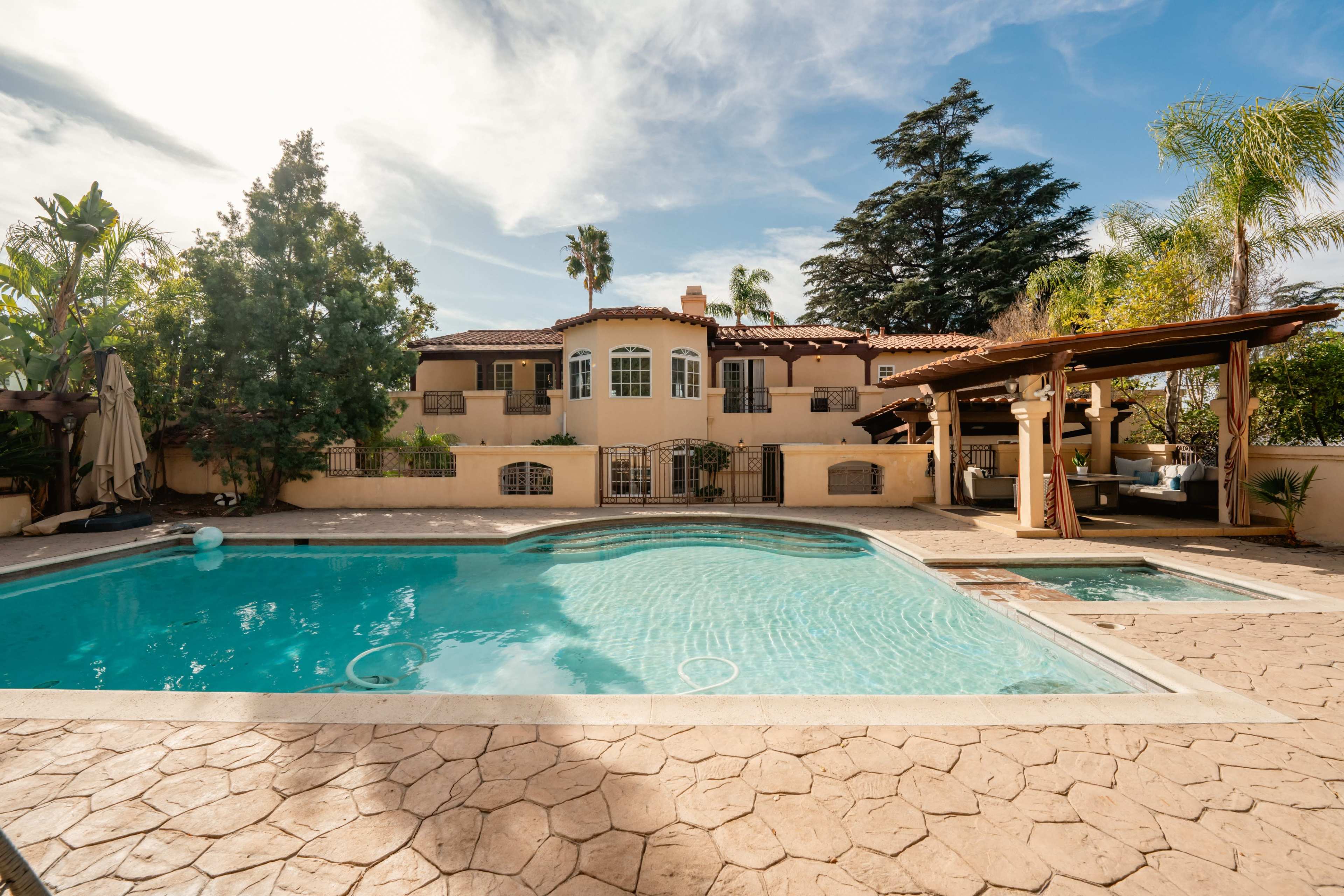 The image shows a large swimming pool surrounded by a patio and a multi-story house with Spanish architectural features, set amid palm trees and other greenery.
