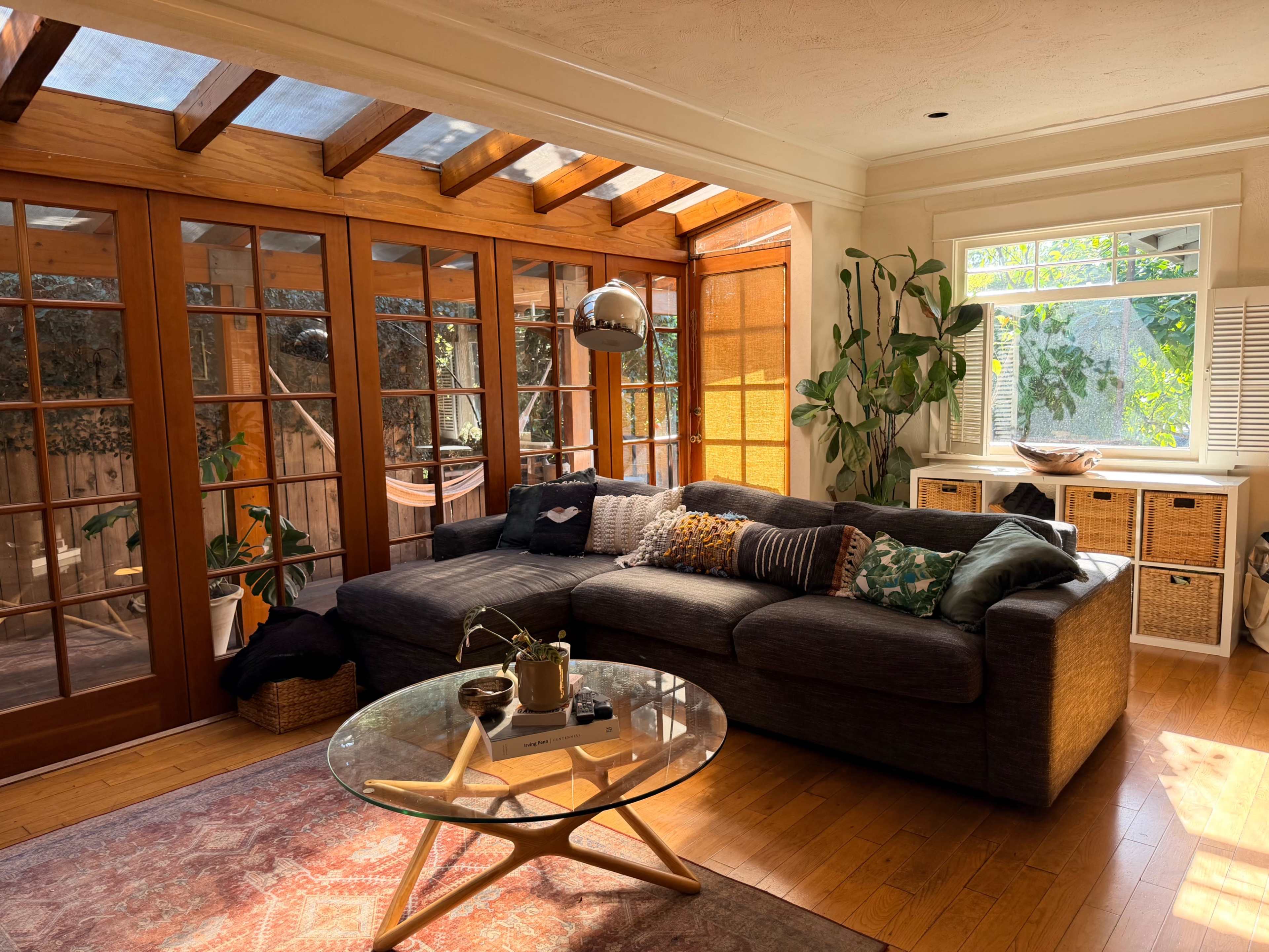 The image shows a sunlit living room featuring a large sectional couch, a circular glass coffee table, and large windows with wooden frames.