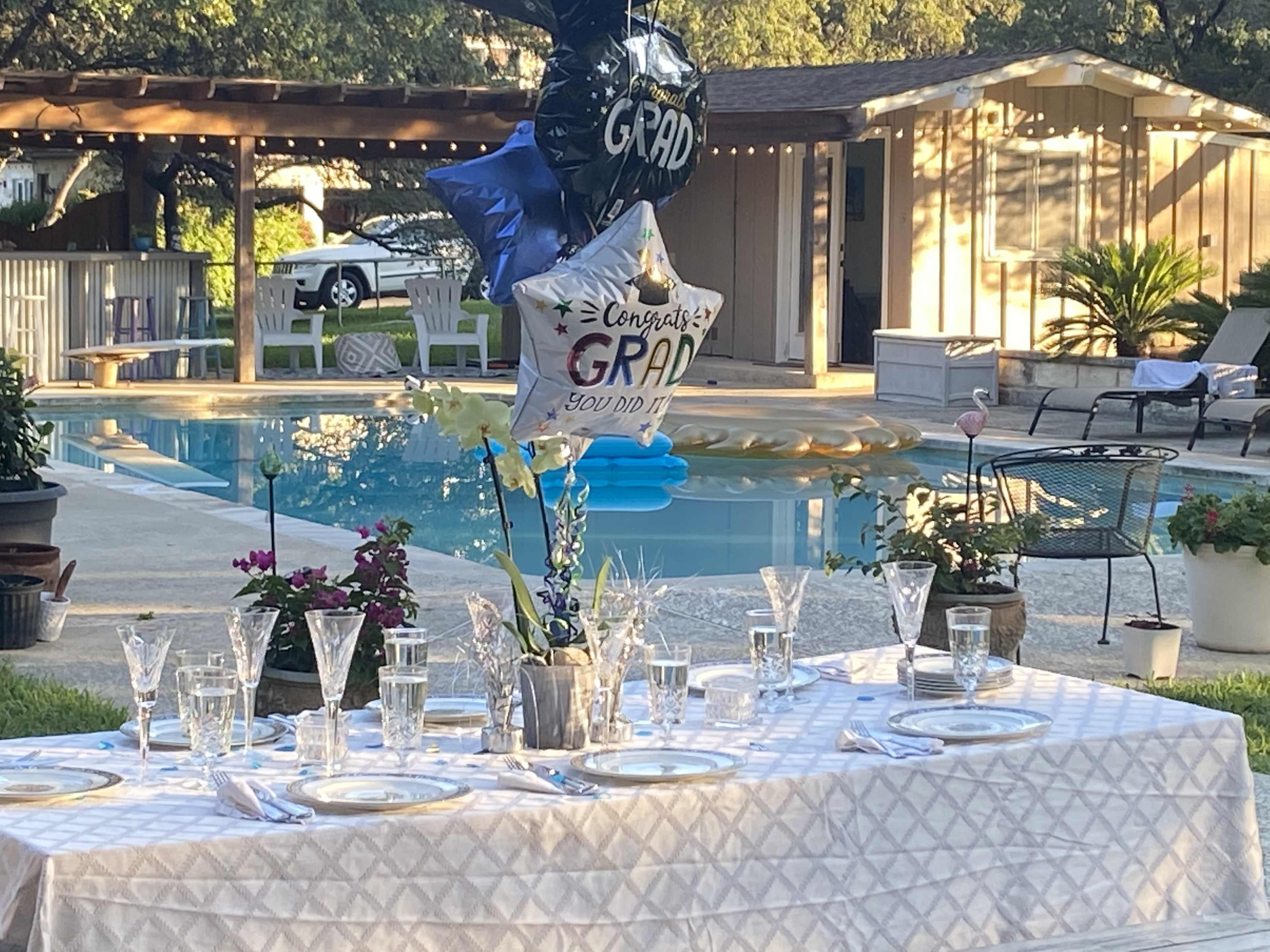 A decorated table is set for a graduation celebration beside a swimming pool, featuring balloons, flowers, and glassware.