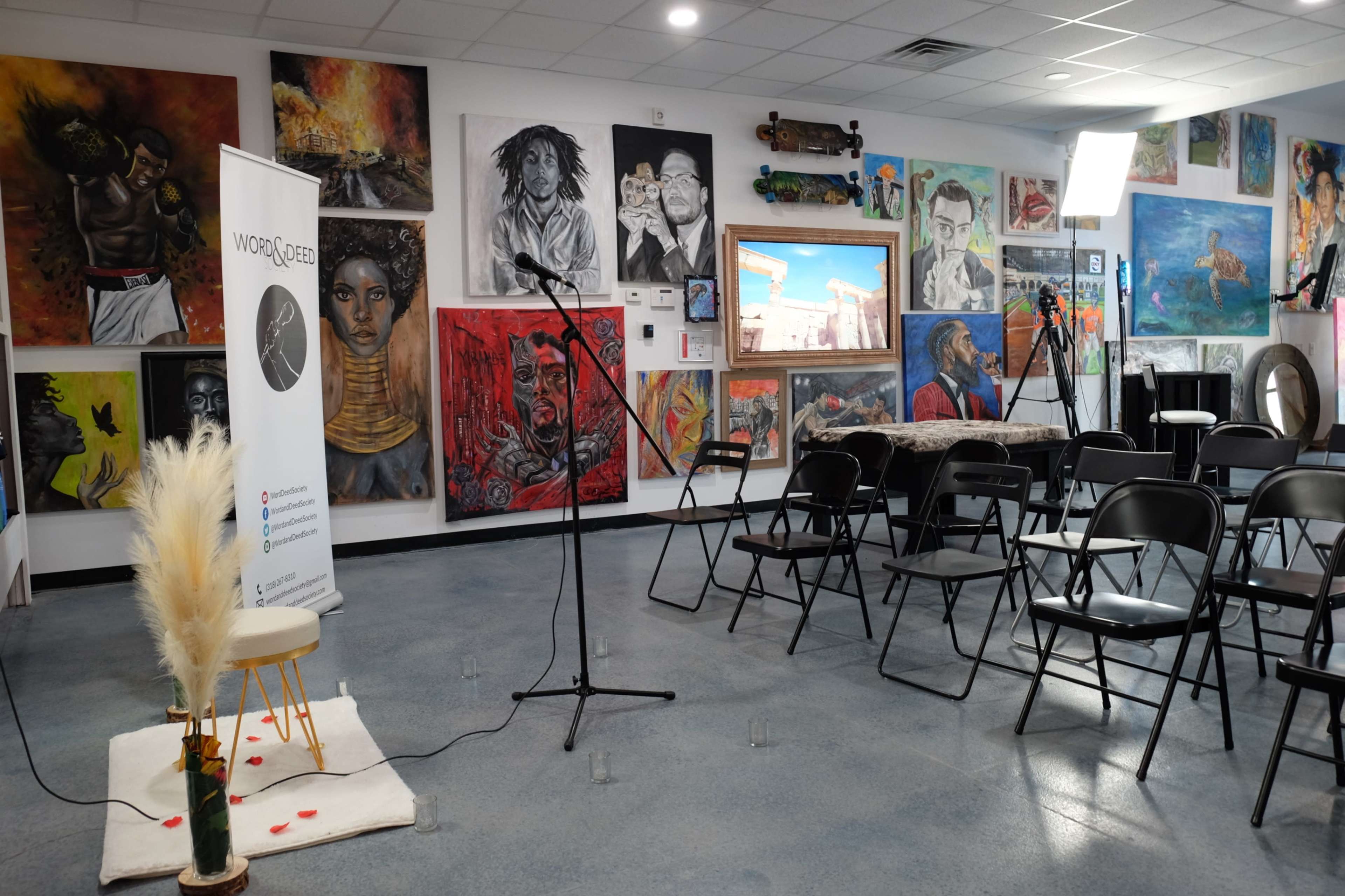 A room set up for an event, featuring empty chairs, a microphone stand, and various colorful artworks on the walls.
