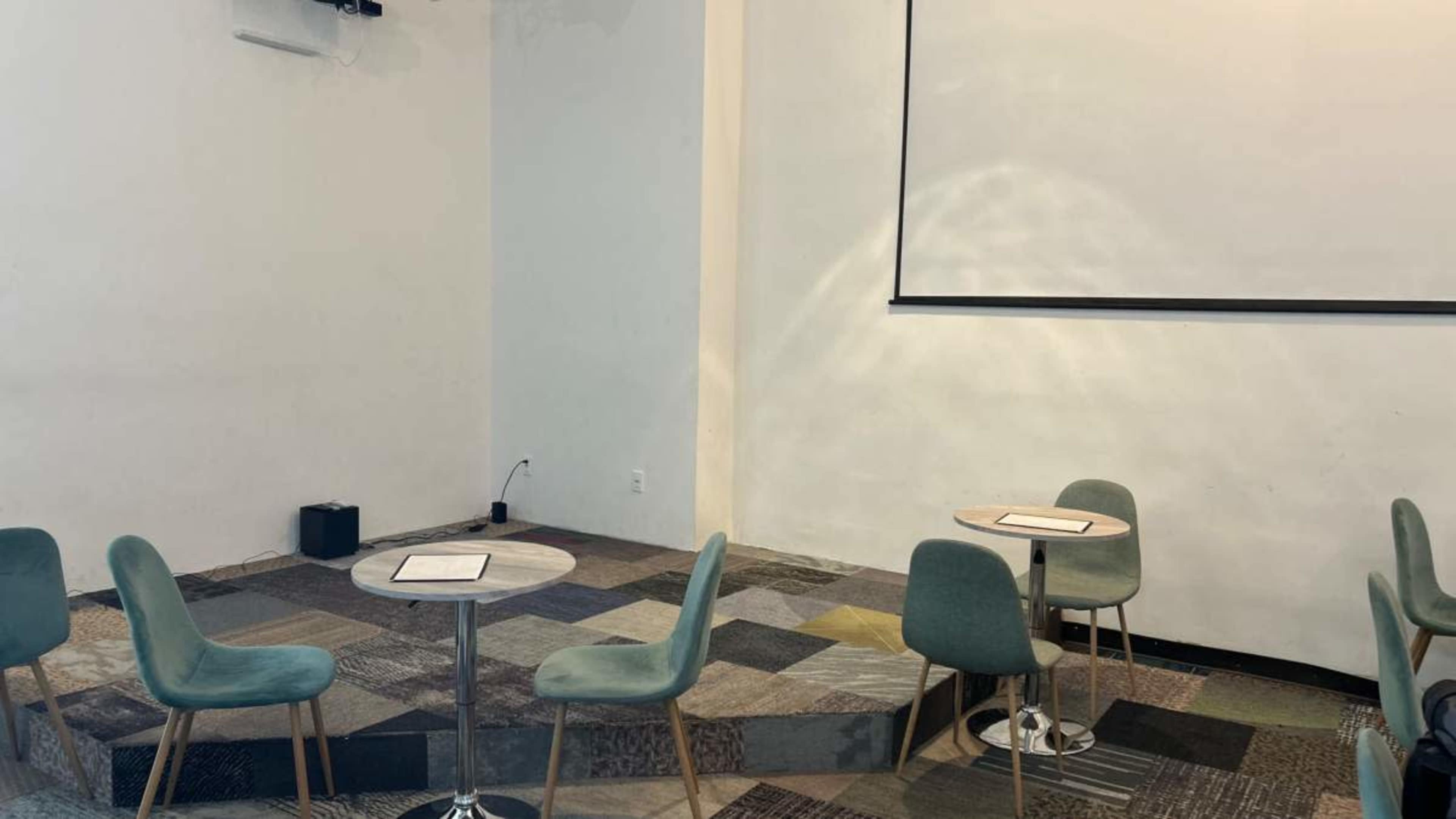 A small, empty meeting space with a presentation screen, several round tables, and green chairs arranged in a circle on patterned flooring.