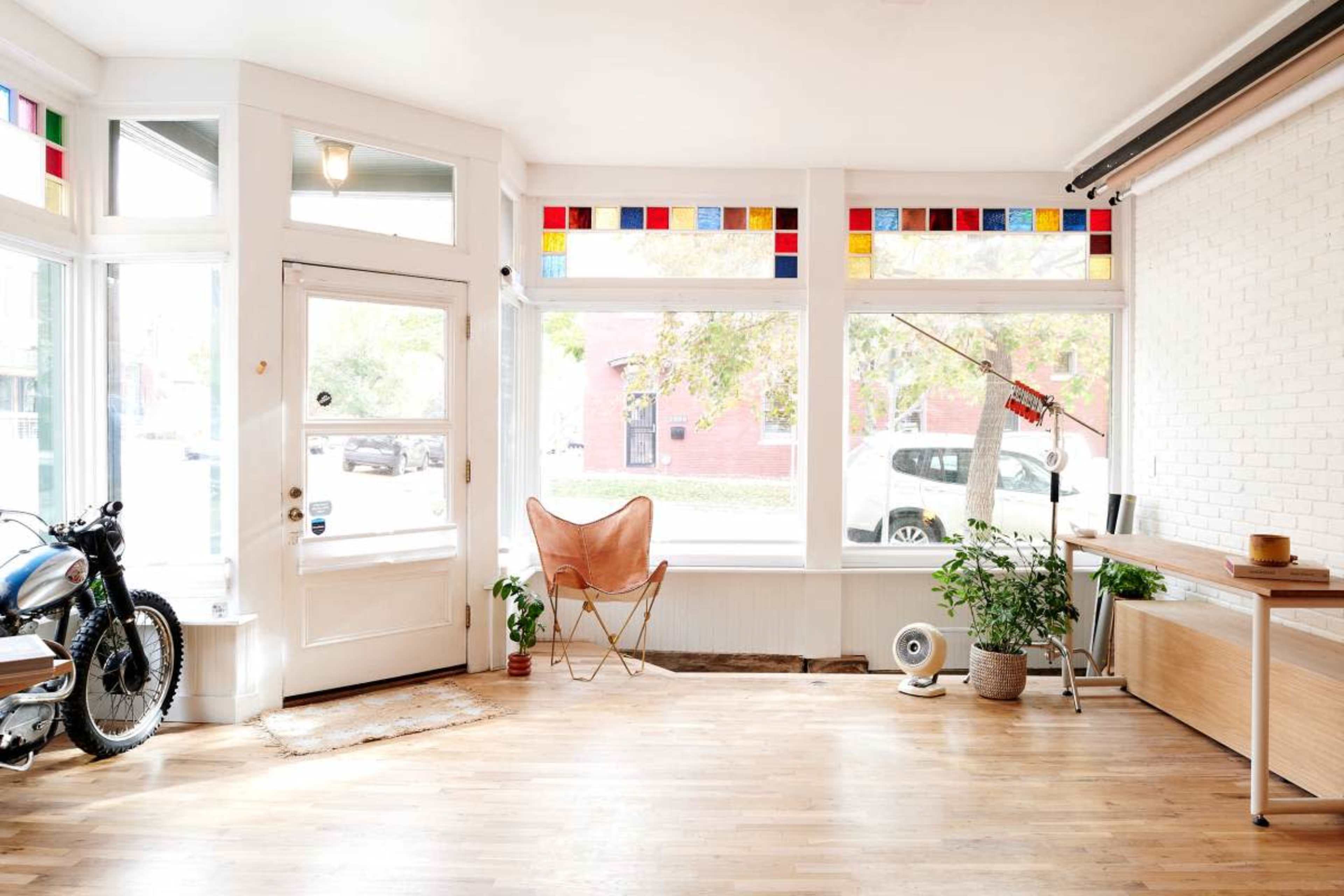 A bright, airy interior space featuring large windows with colored panes, a decorative chair, a motorcycle parked inside, and a wooden table.