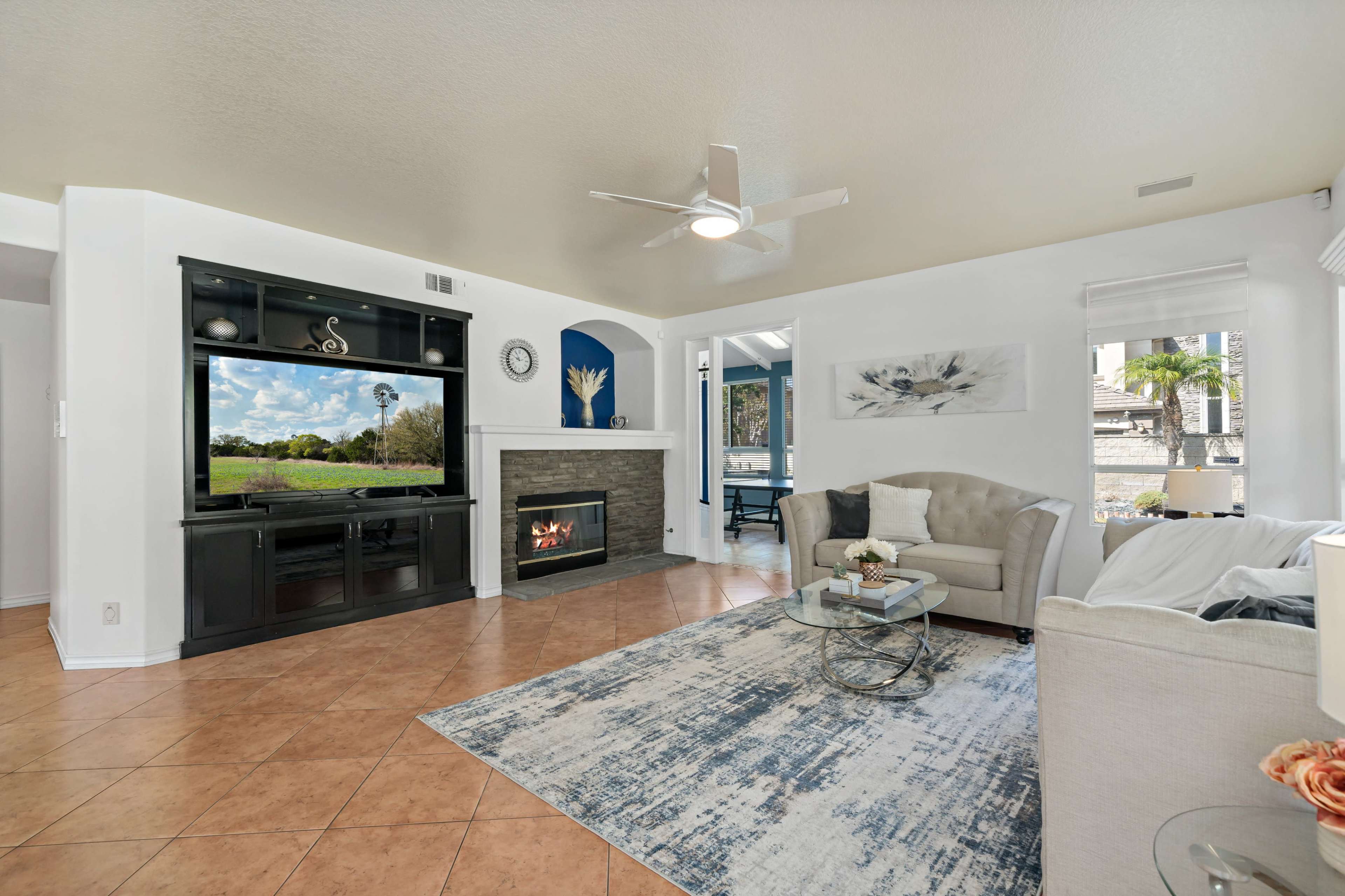 A living room features a large television above a fireplace, a light-colored sofa, a round coffee table, and a textured rug, with a view of the outdoors visible through a window.