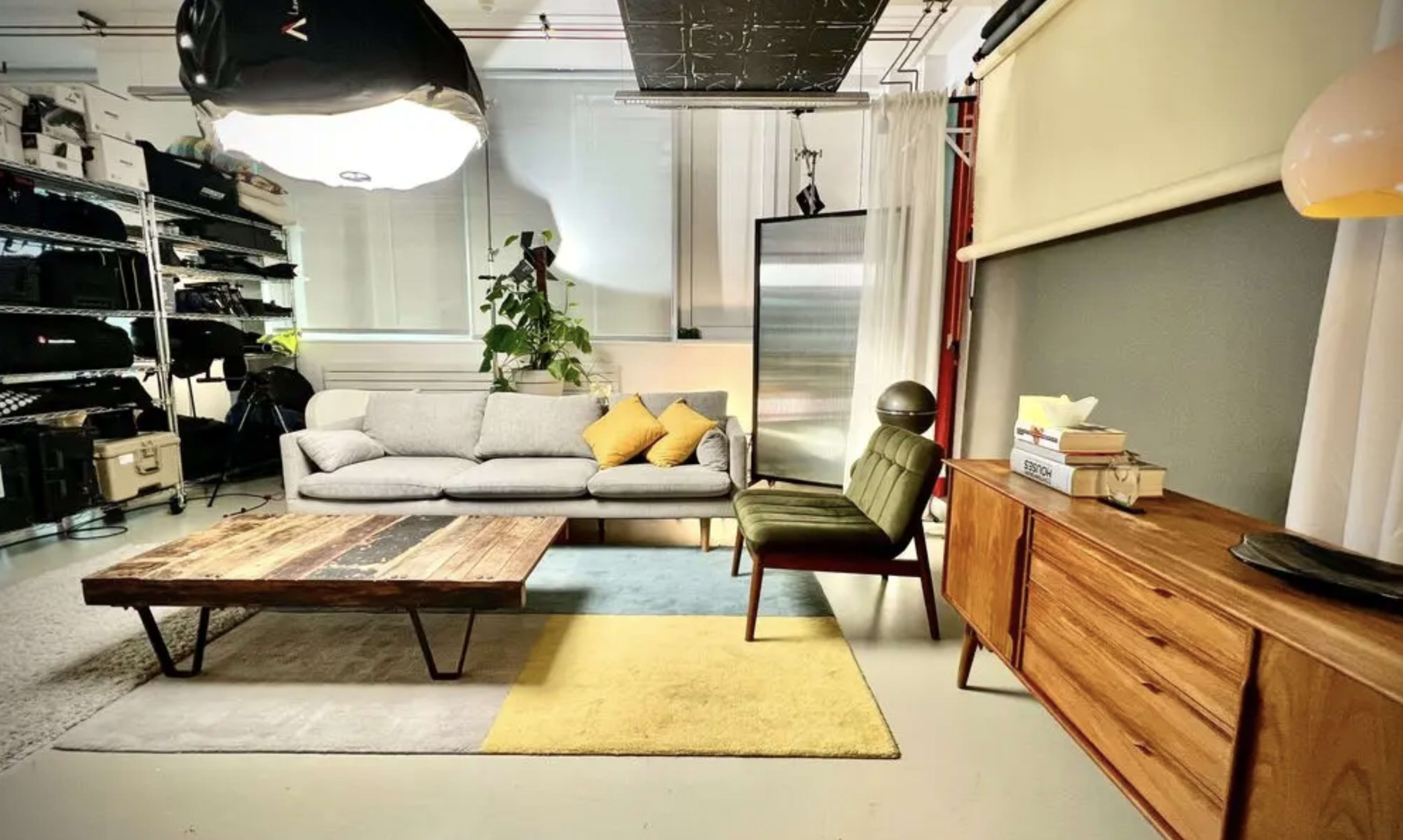 The image features a modern, well-lit room with a gray sofa, a green armchair, a wooden coffee table, and a mid-century style sideboard, surrounded by various studio equipment and storage shelves.