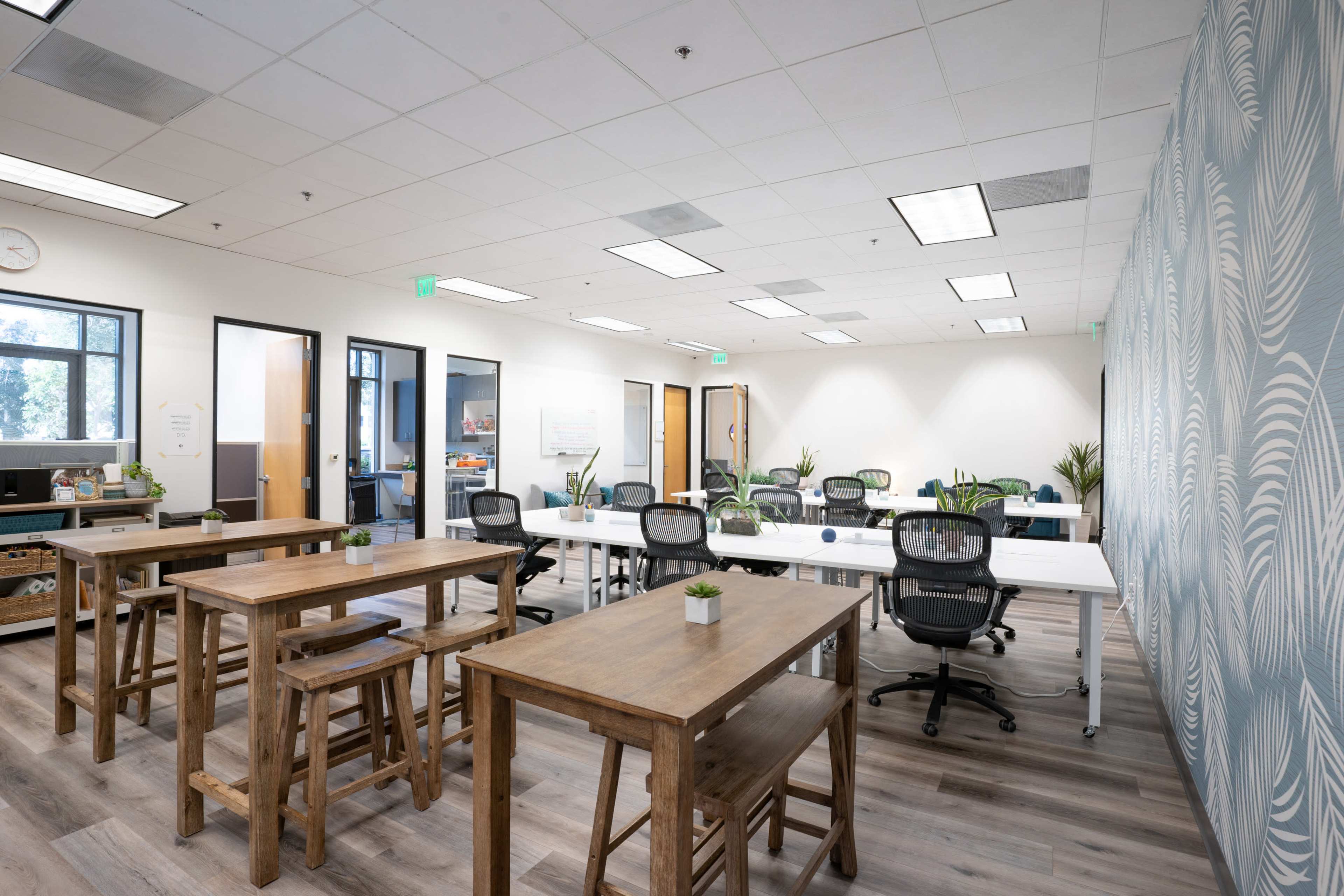 The image shows a modern office space with several wooden tables, ergonomic chairs, and plants, featuring large windows that allow natural light to enter.