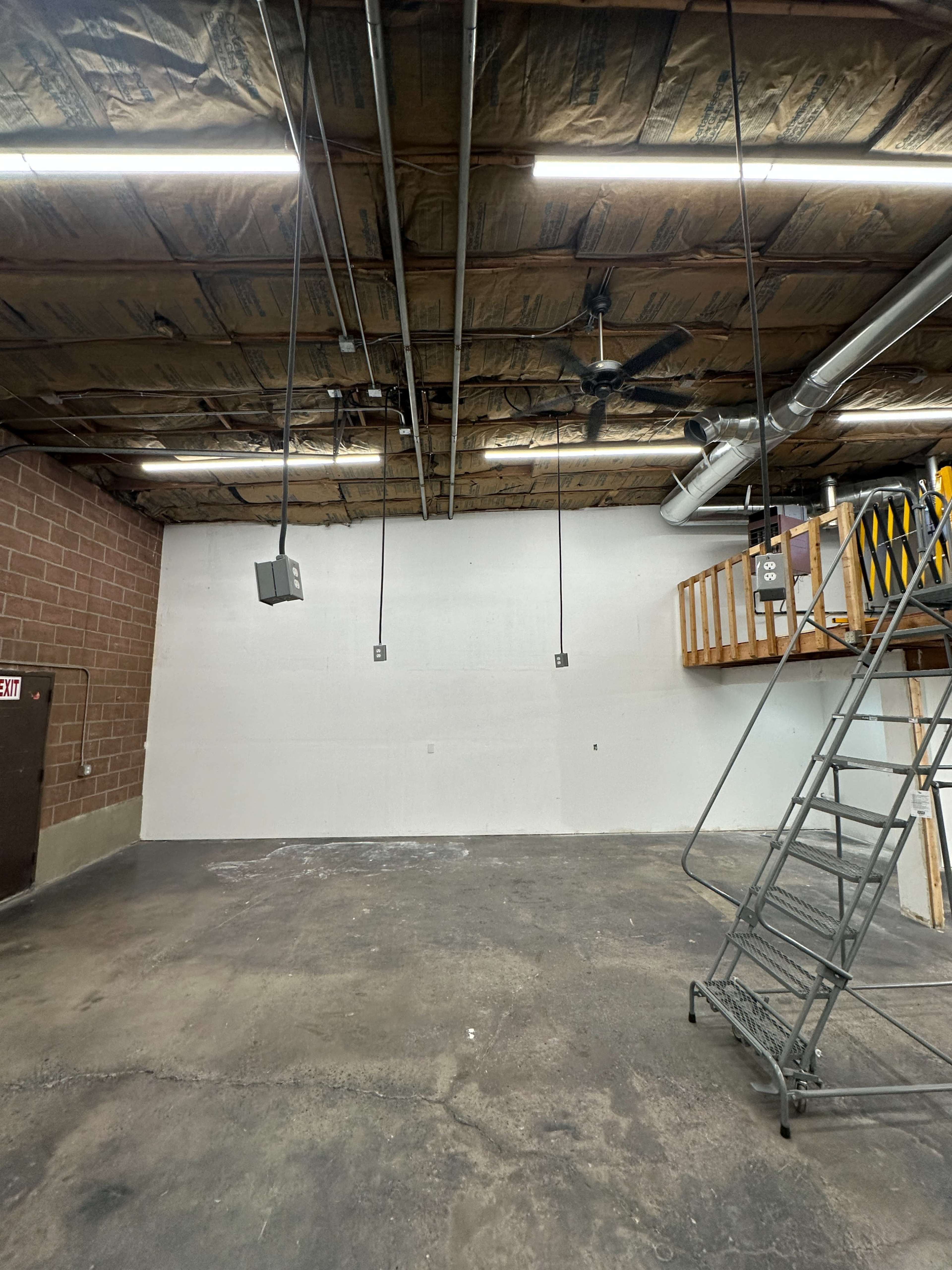 The image shows an empty industrial space with exposed ceiling beams, overhead lighting, and a sliding ladder against one wall.