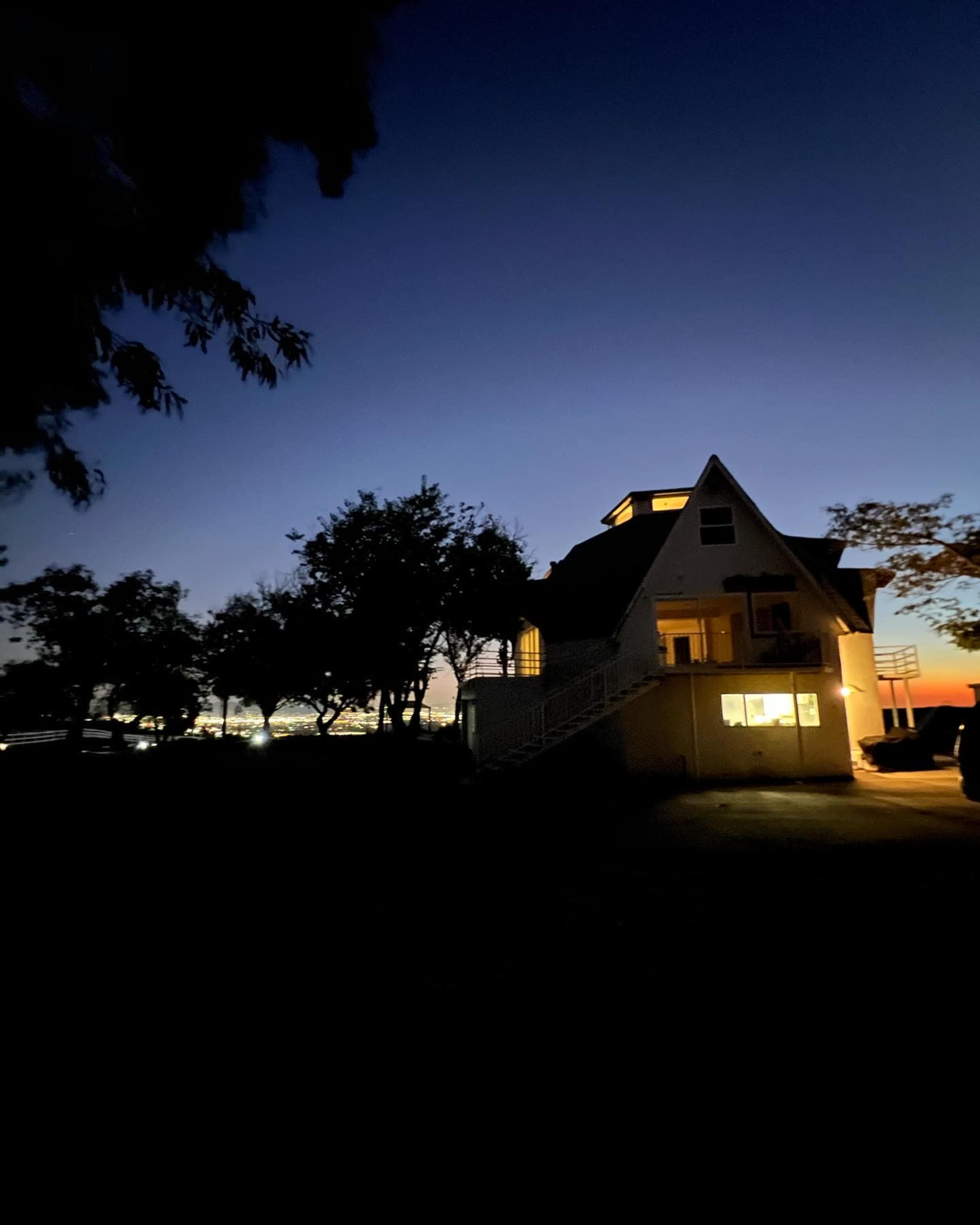 A house with a triangular roof is illuminated at dusk, surrounded by trees against a darkening sky.