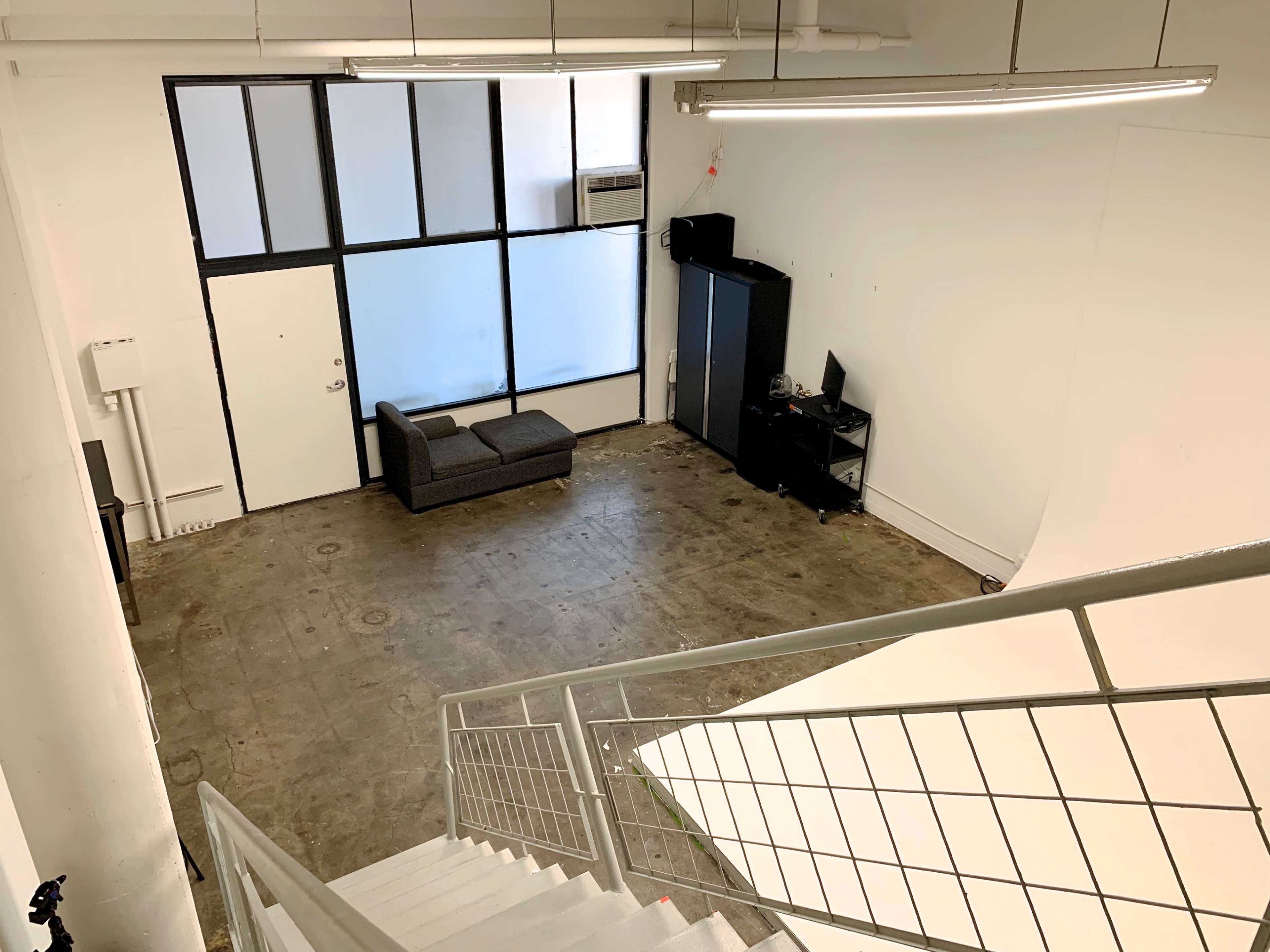 A spacious, empty room with large windows, a gray couch, a desk with a computer, and a staircase leading down to a lower level.