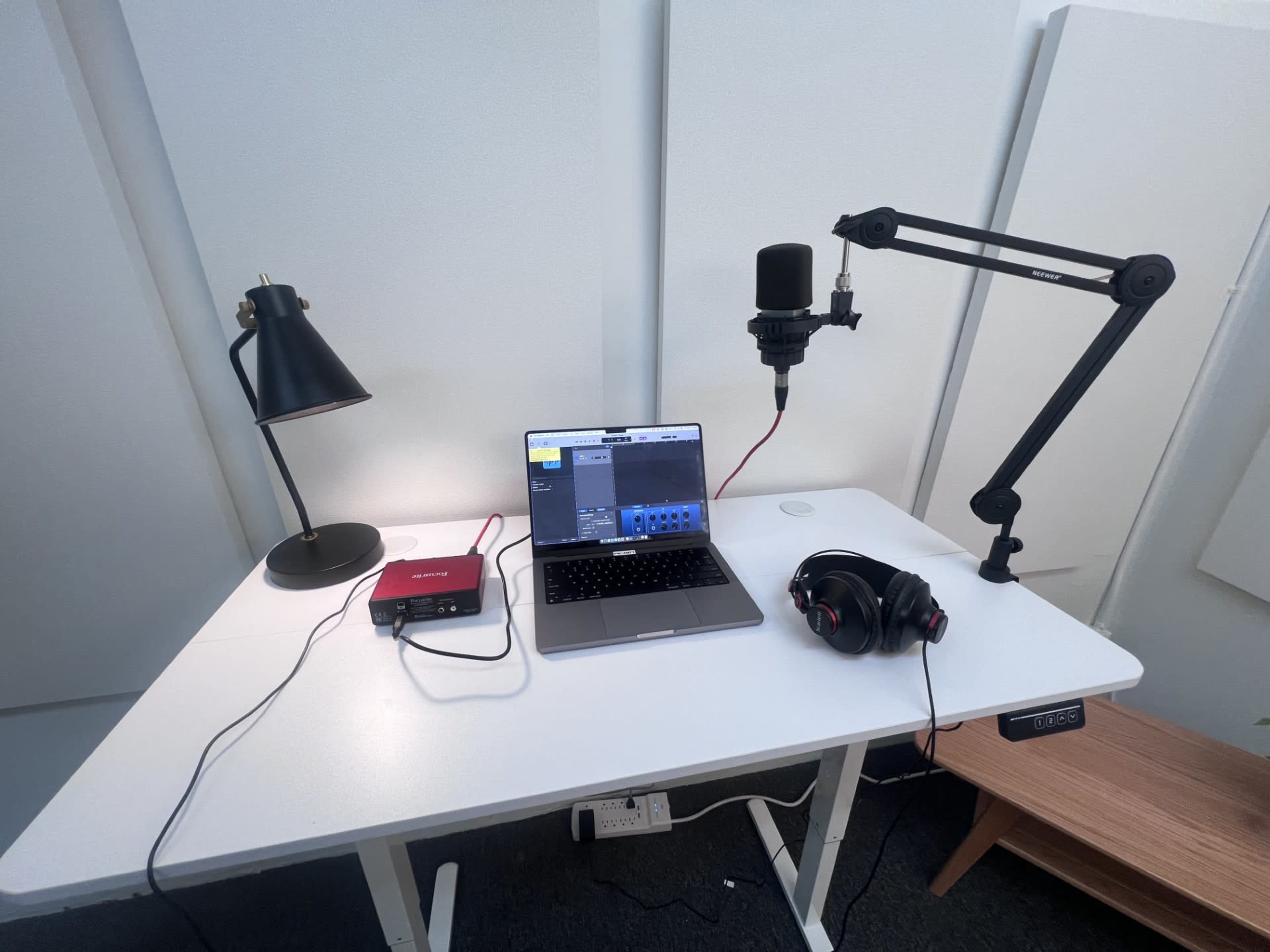 The image shows a desk setup with a laptop, microphone on an arm, headphones, an audio interface, and a lamp in a soundproof environment.