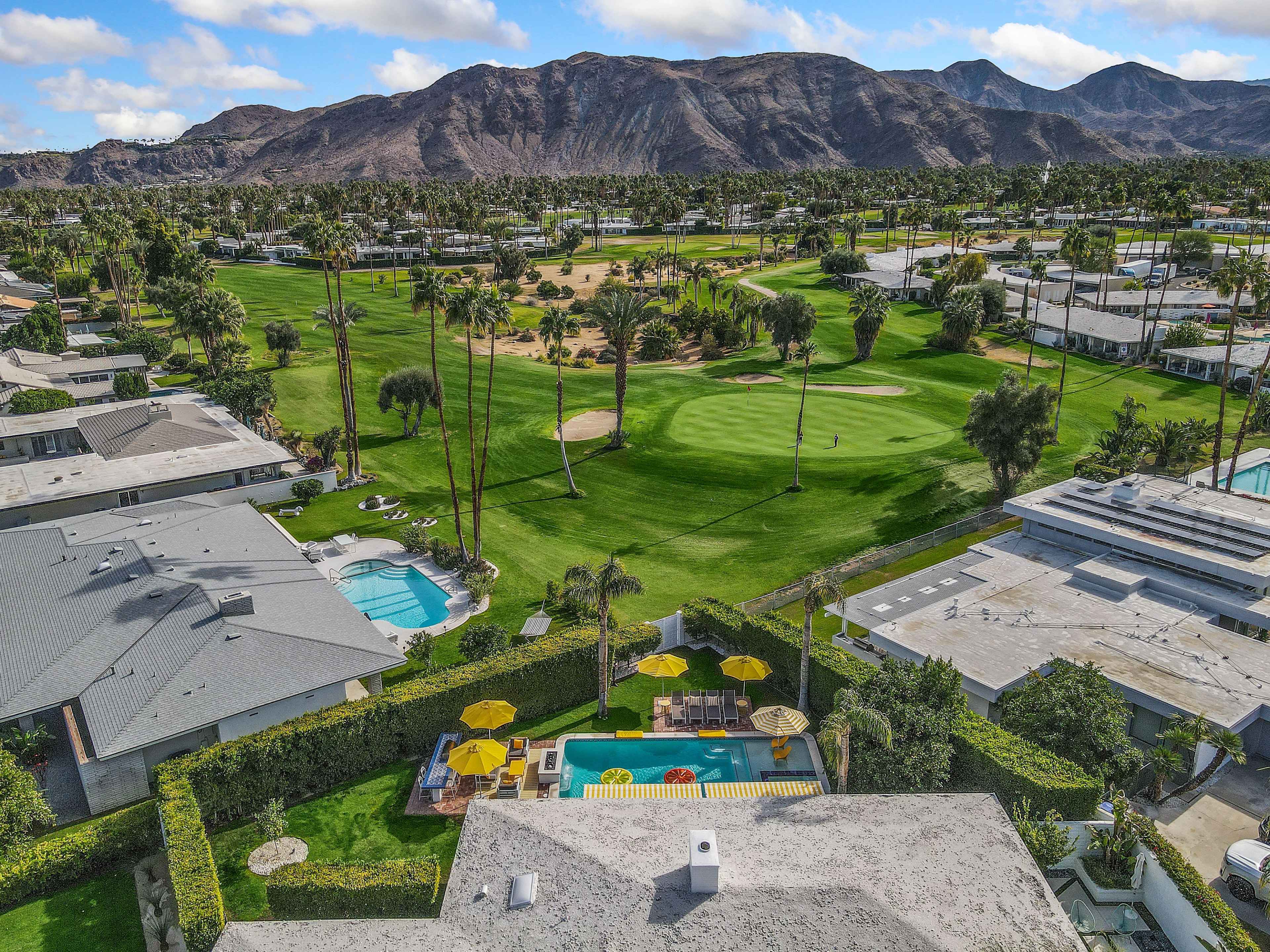 The image shows a residential area with houses, a swimming pool, and a golf course, surrounded by palm trees and mountains in the background.