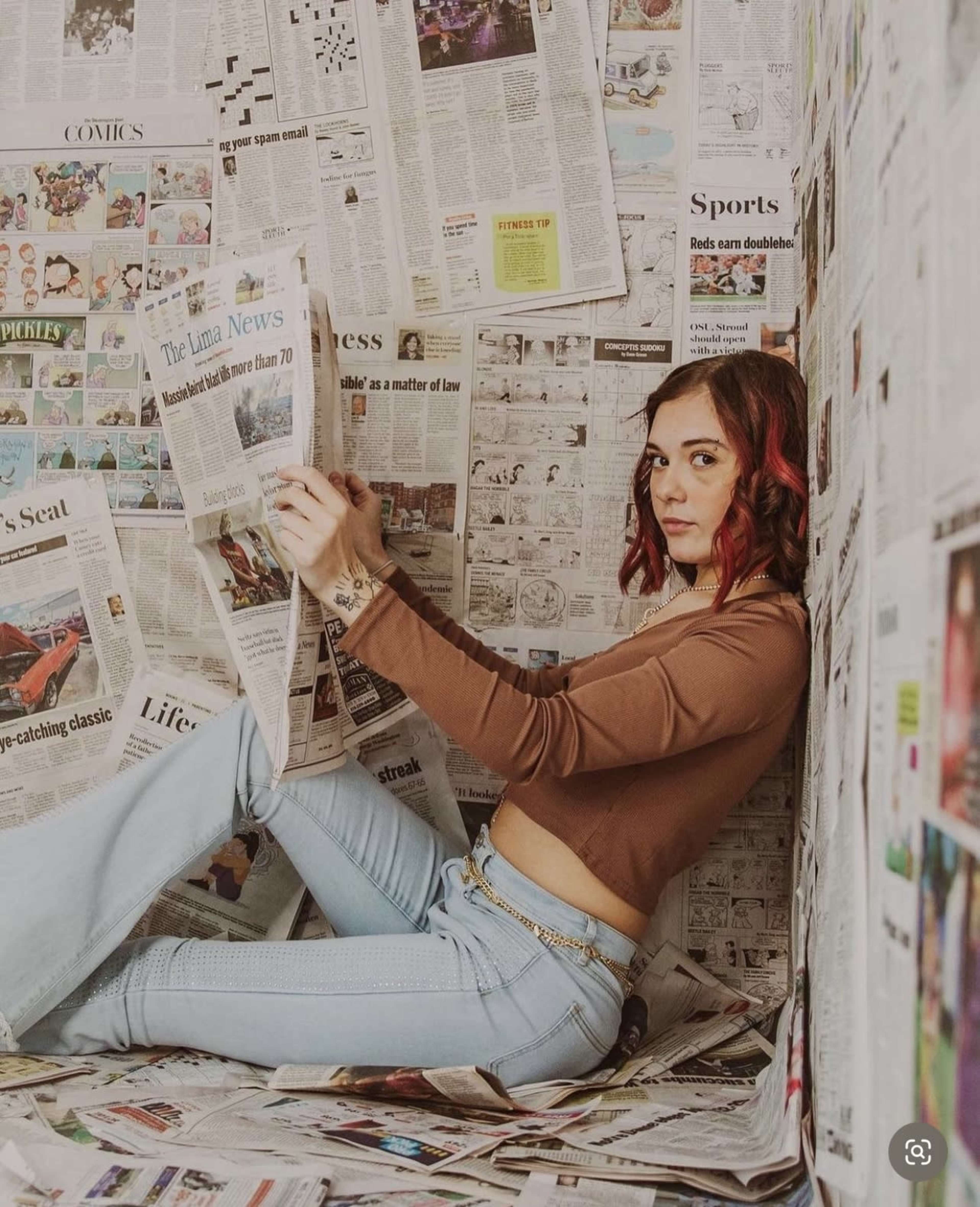 A woman sits in a corner surrounded by walls made of newspaper, holding a piece of newspaper in her hands.