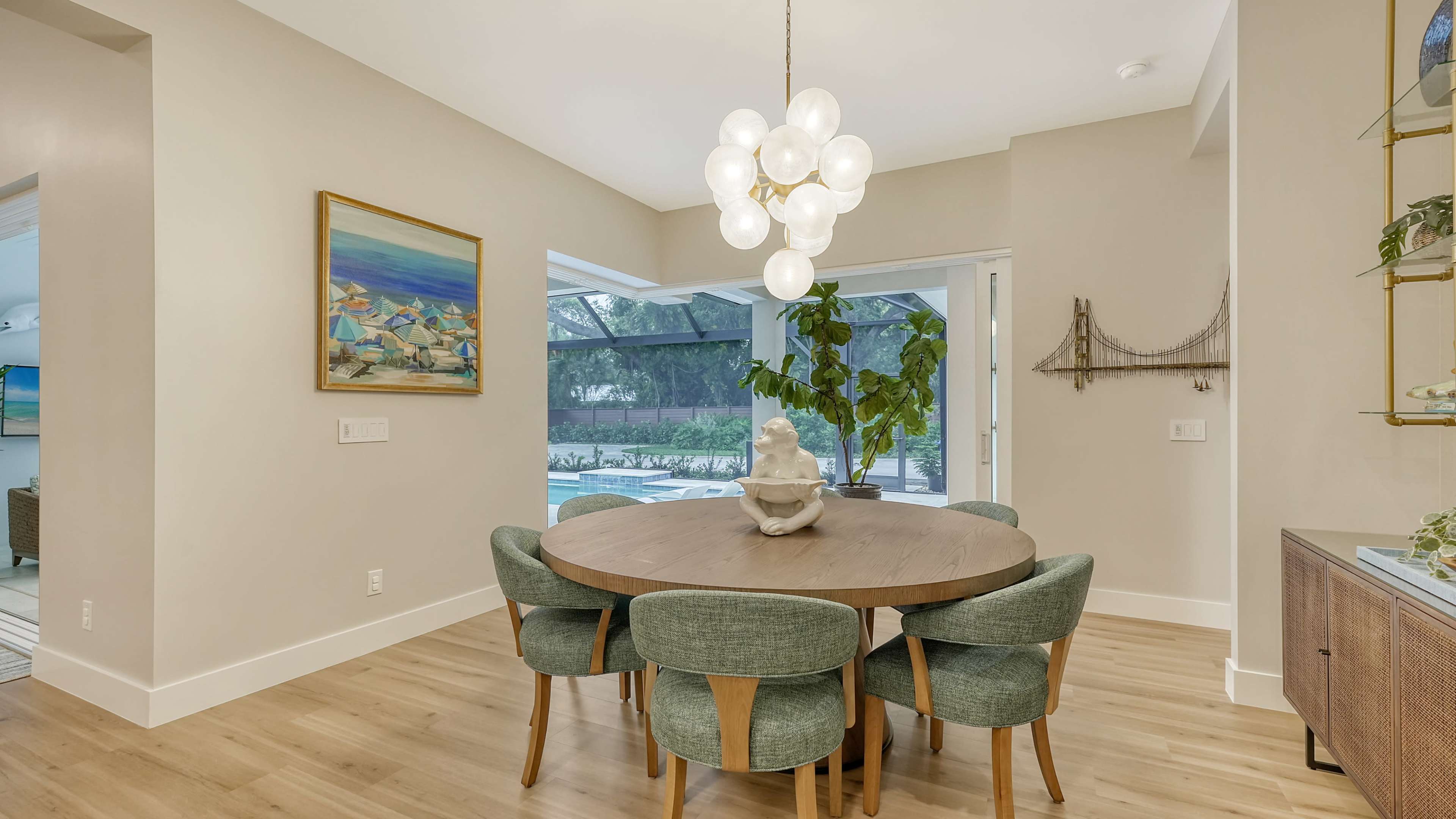 A round wooden dining table surrounded by upholstered chairs is positioned near a large window with a view of a patio and pool area.