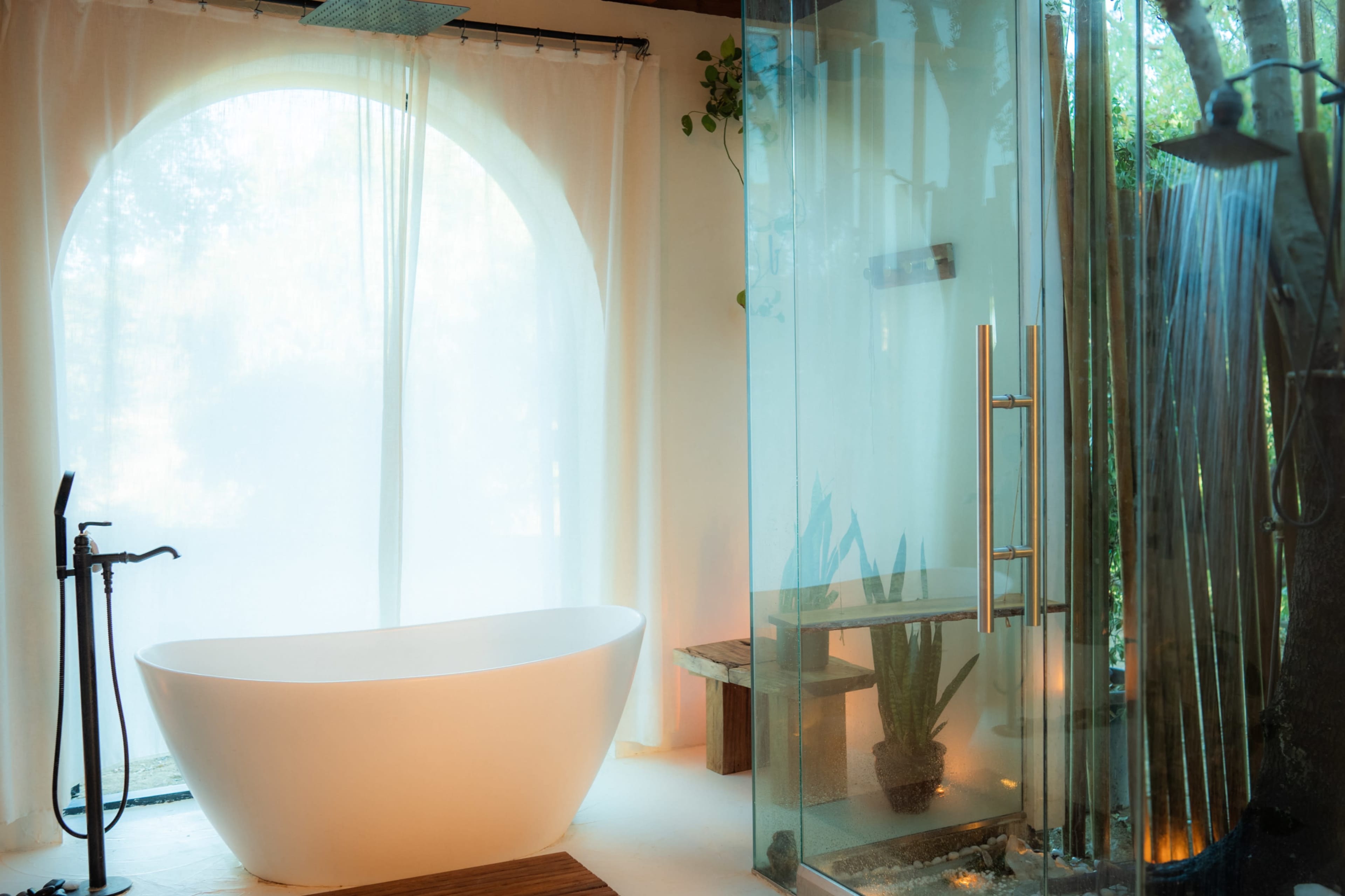 The image shows a modern bathroom with a freestanding white bathtub, a glass-enclosed shower, and natural light filtering through sheer curtains.