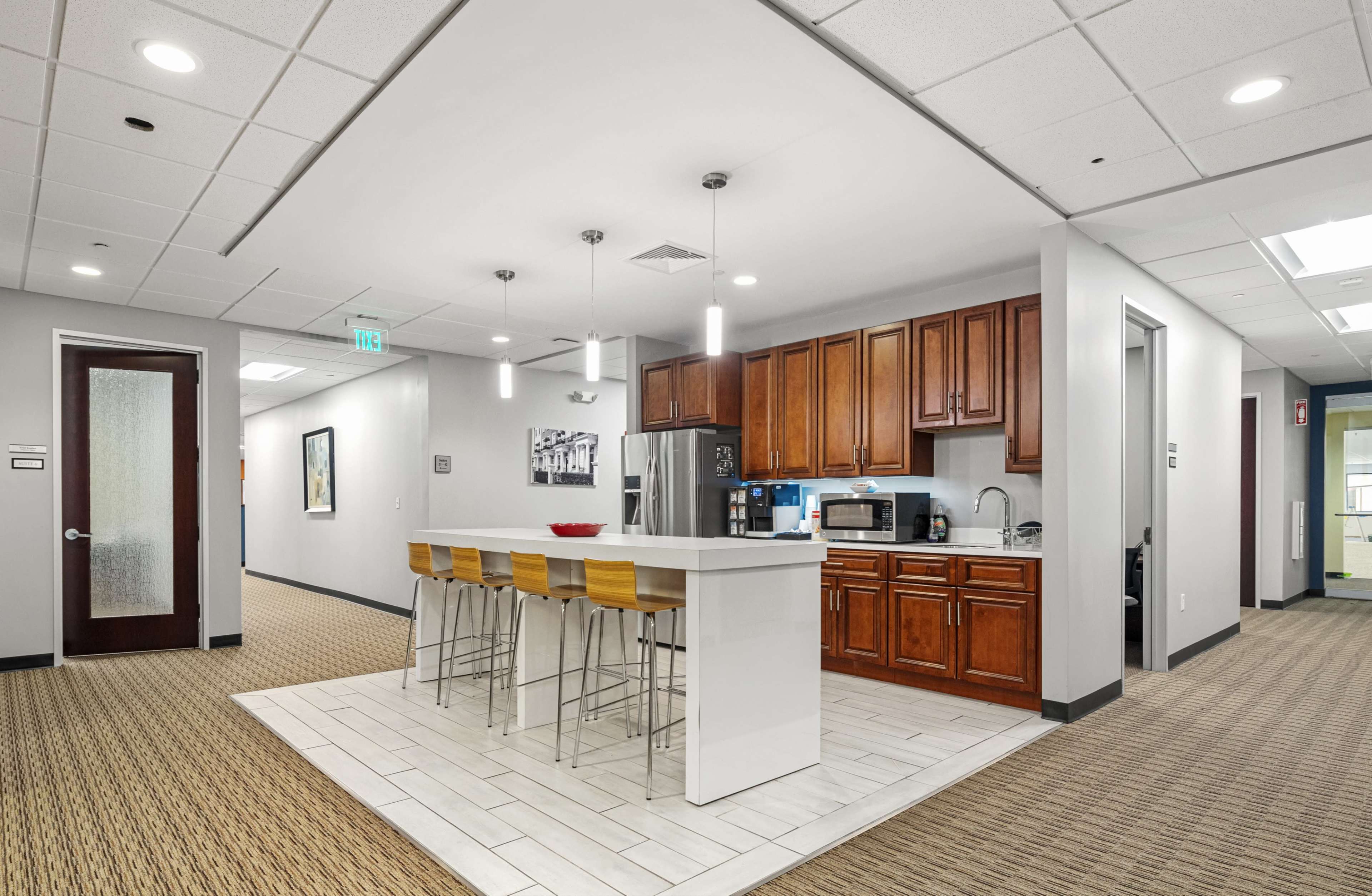 A modern kitchen area with a bar countertop and high stools, featuring wooden cabinets, a refrigerator, and a microwave.