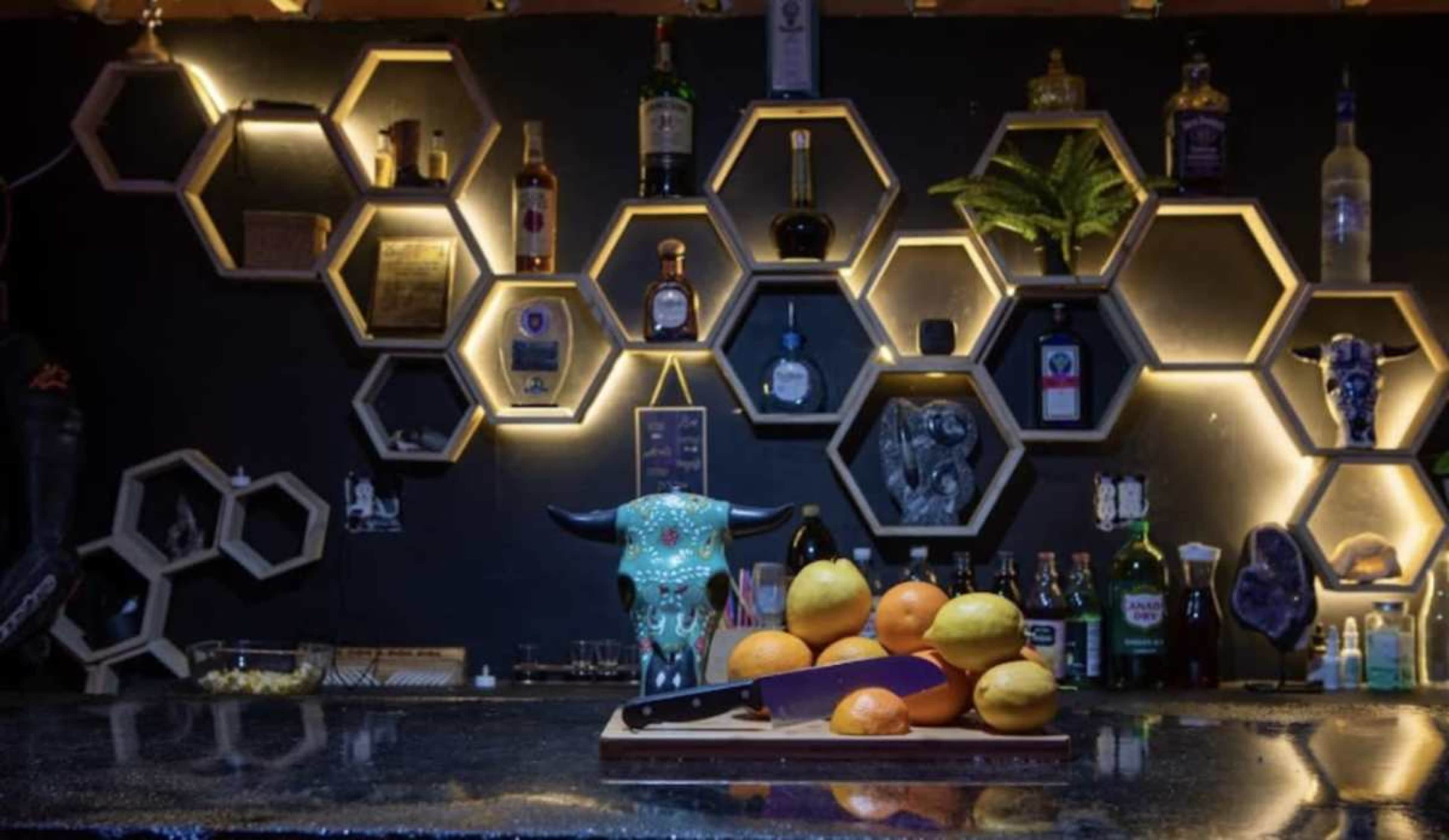 A bar countertop displays a wooden cutting board with sliced fruits, while a wall featuring hexagonal shelves holds various bottles and decorative items.
