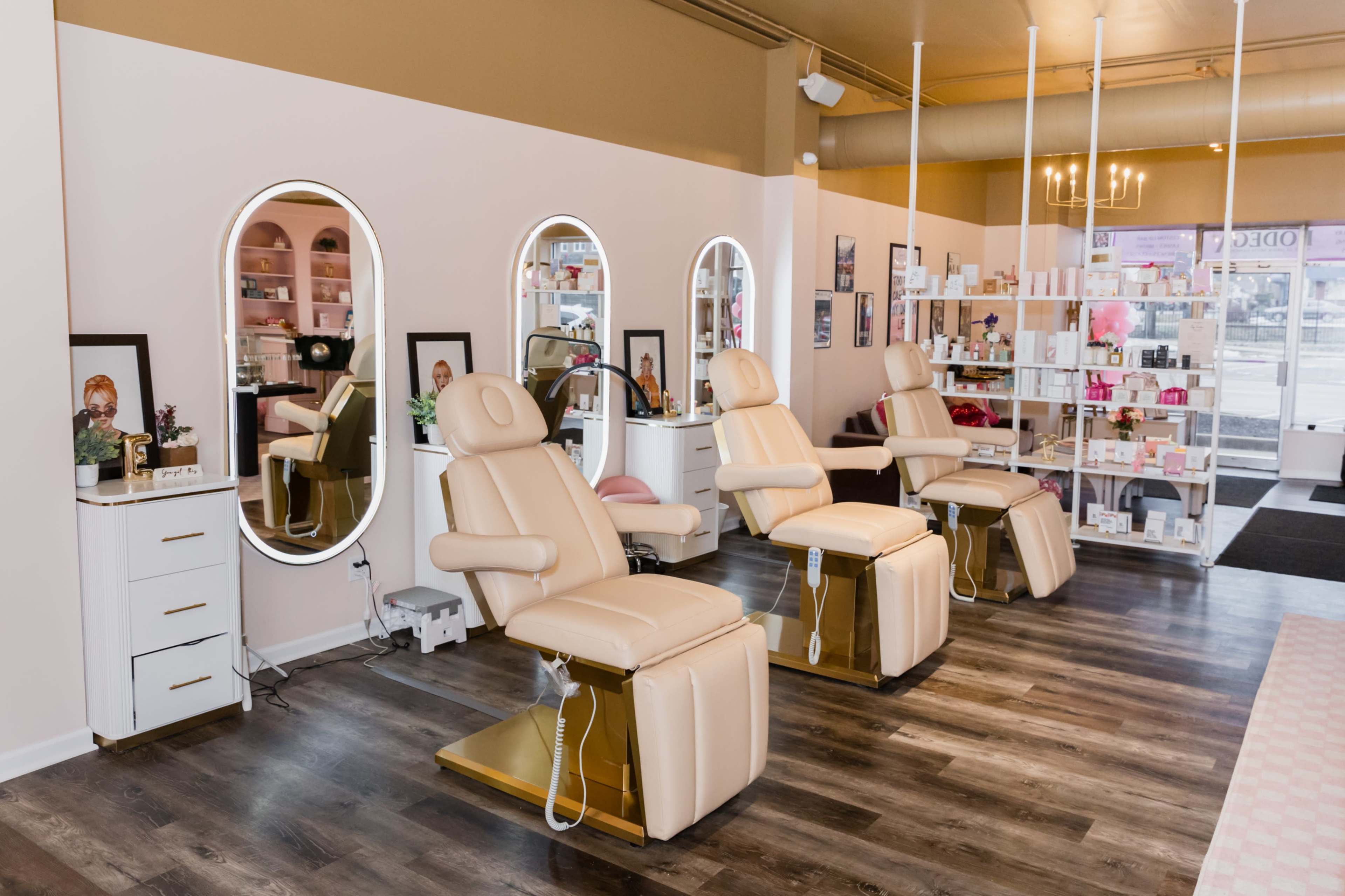 A modern beauty salon with three beige treatment chairs arranged in front of illuminated mirrors against a backdrop of shelves filled with assorted beauty products.