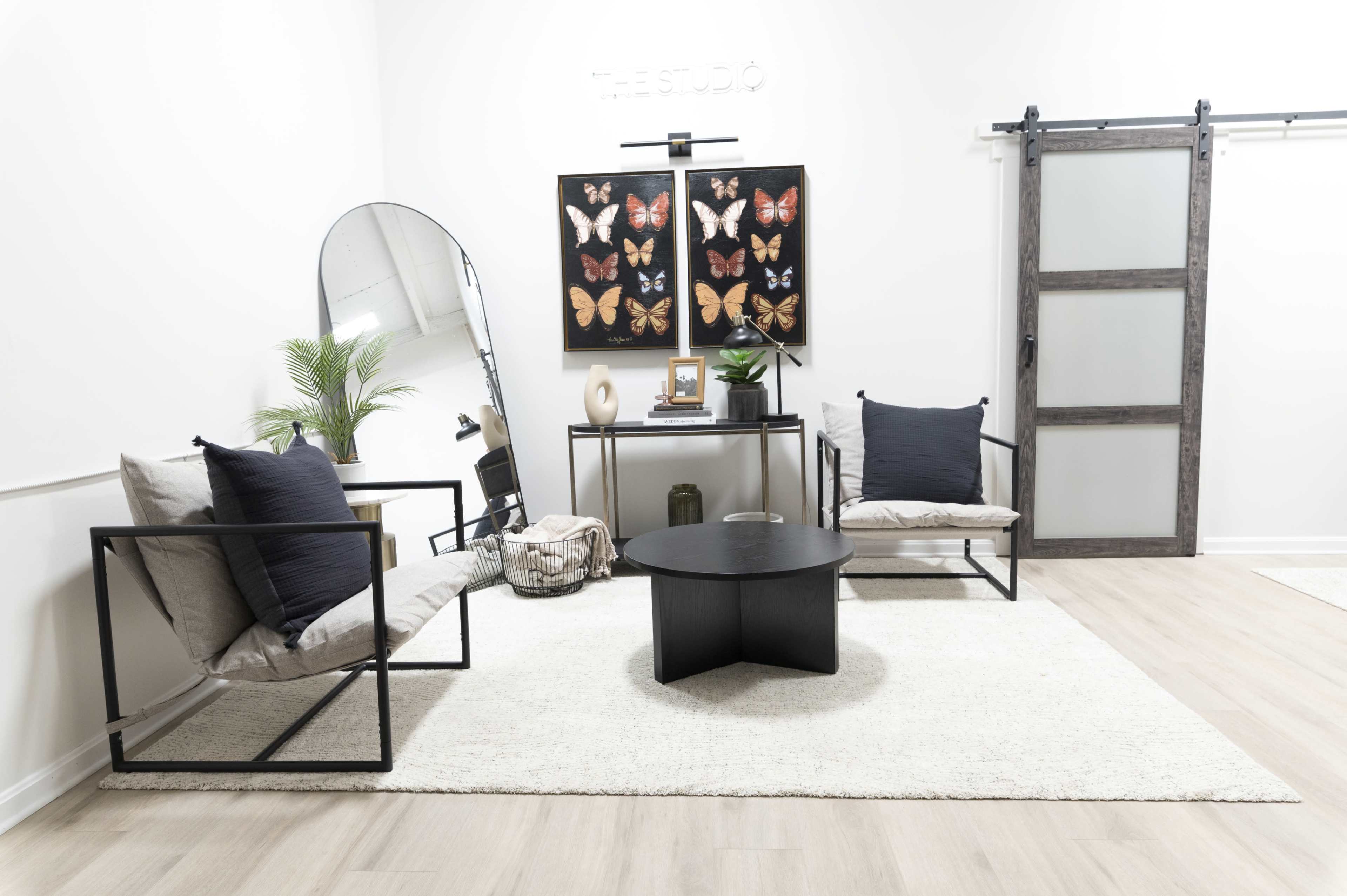 The room features two metal-framed chairs with cushions, a round black coffee table, a wall-mounted butterfly art piece, and a sliding door, all arranged on a light-colored rug.