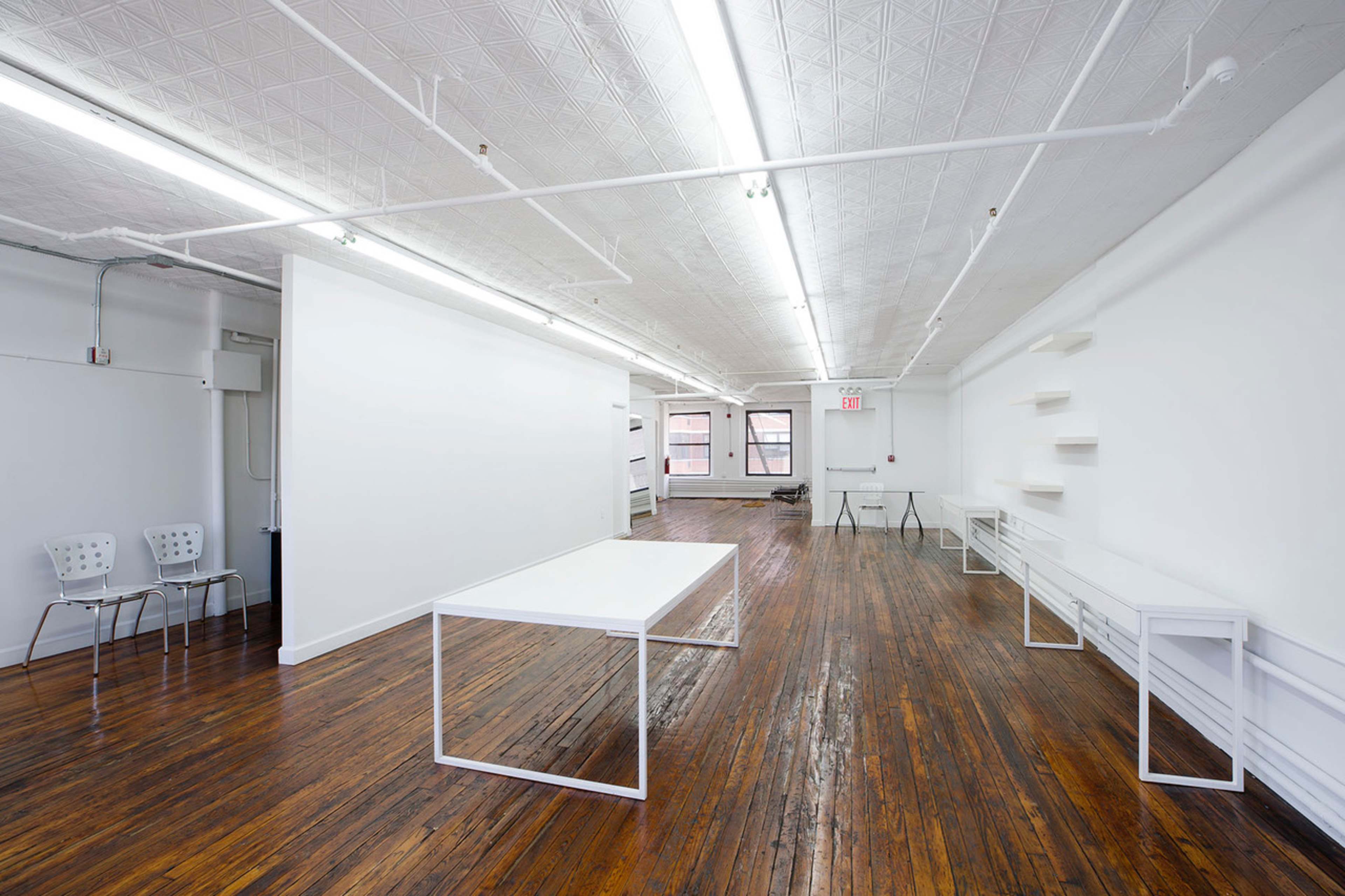 A spacious, well-lit room features wooden floors and minimal furnishings, including a white table and several chairs against the walls.