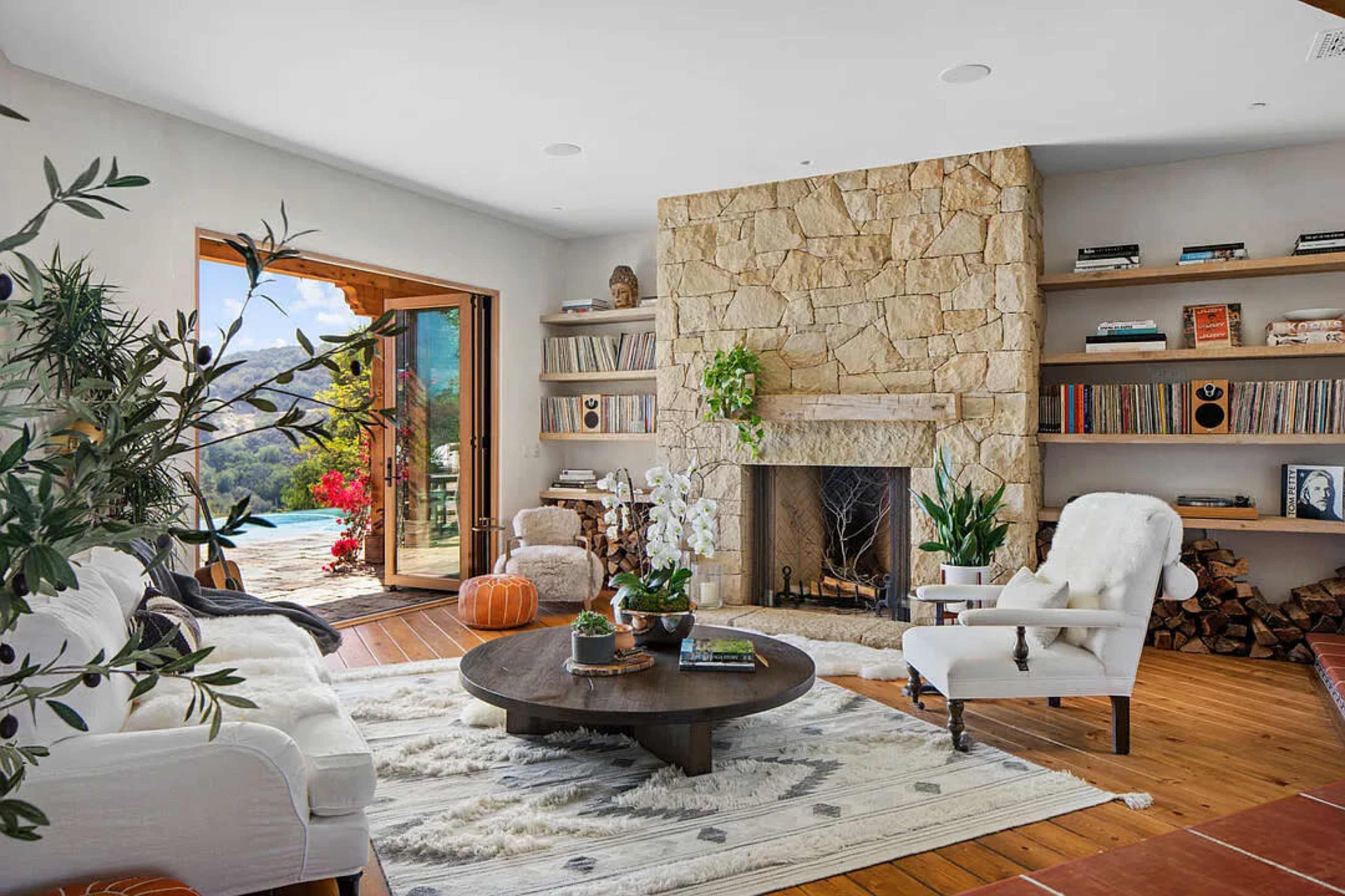 The image shows a modern living room featuring a stone fireplace, a circular coffee table, bookshelves, and large windows that open to a scenic outdoor view.