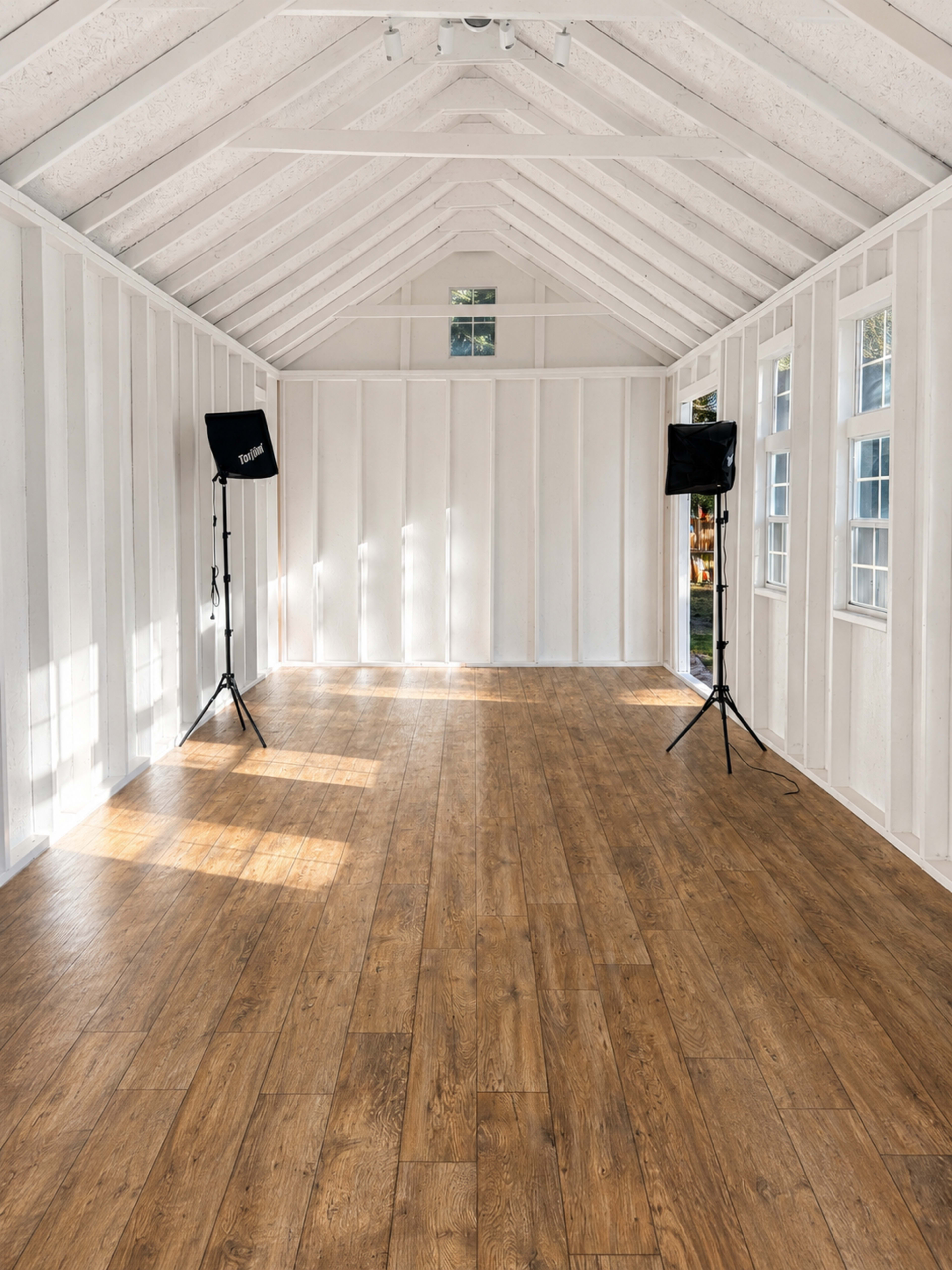 The interior of a bright, empty room features wooden flooring, white walls, and two light stands positioned on either side.