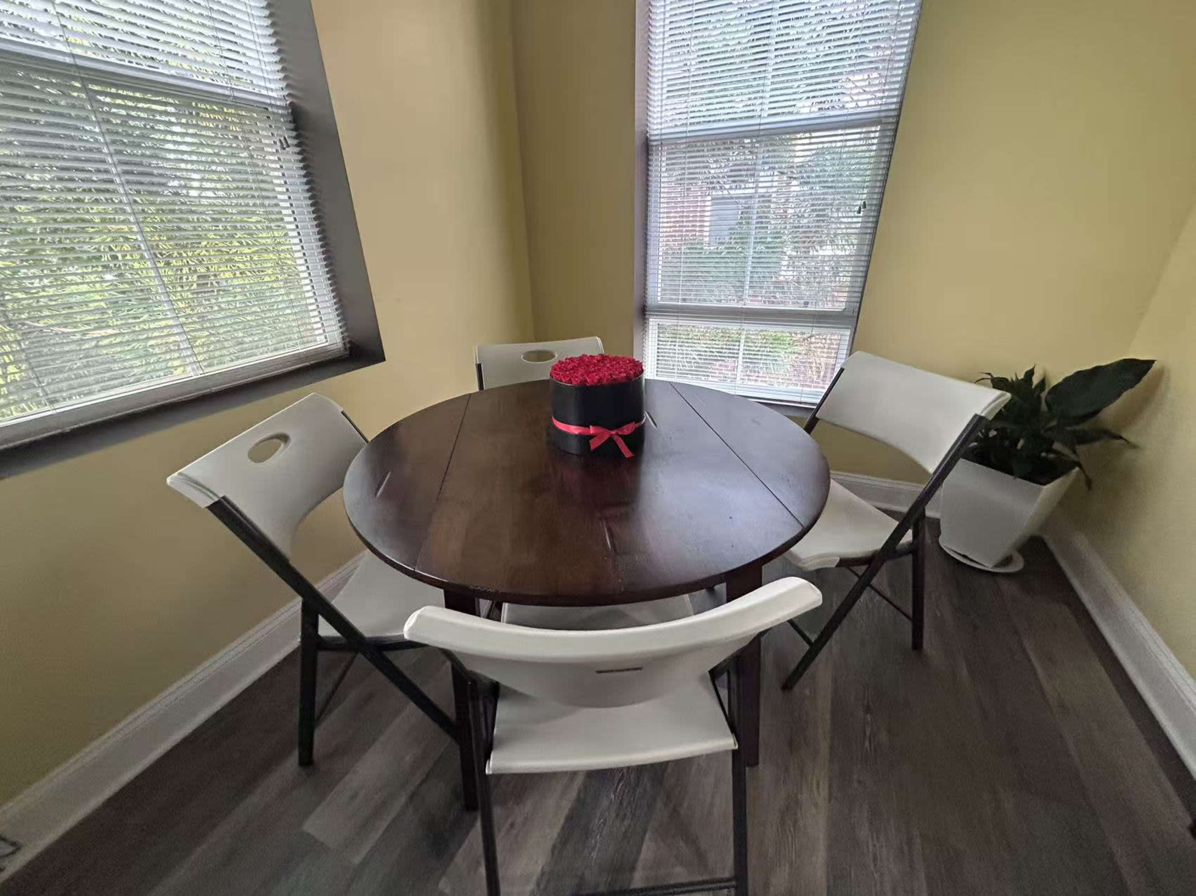 A round wooden table is set in a sunny corner with four white folding chairs and a floral arrangement in a black and red container on top.