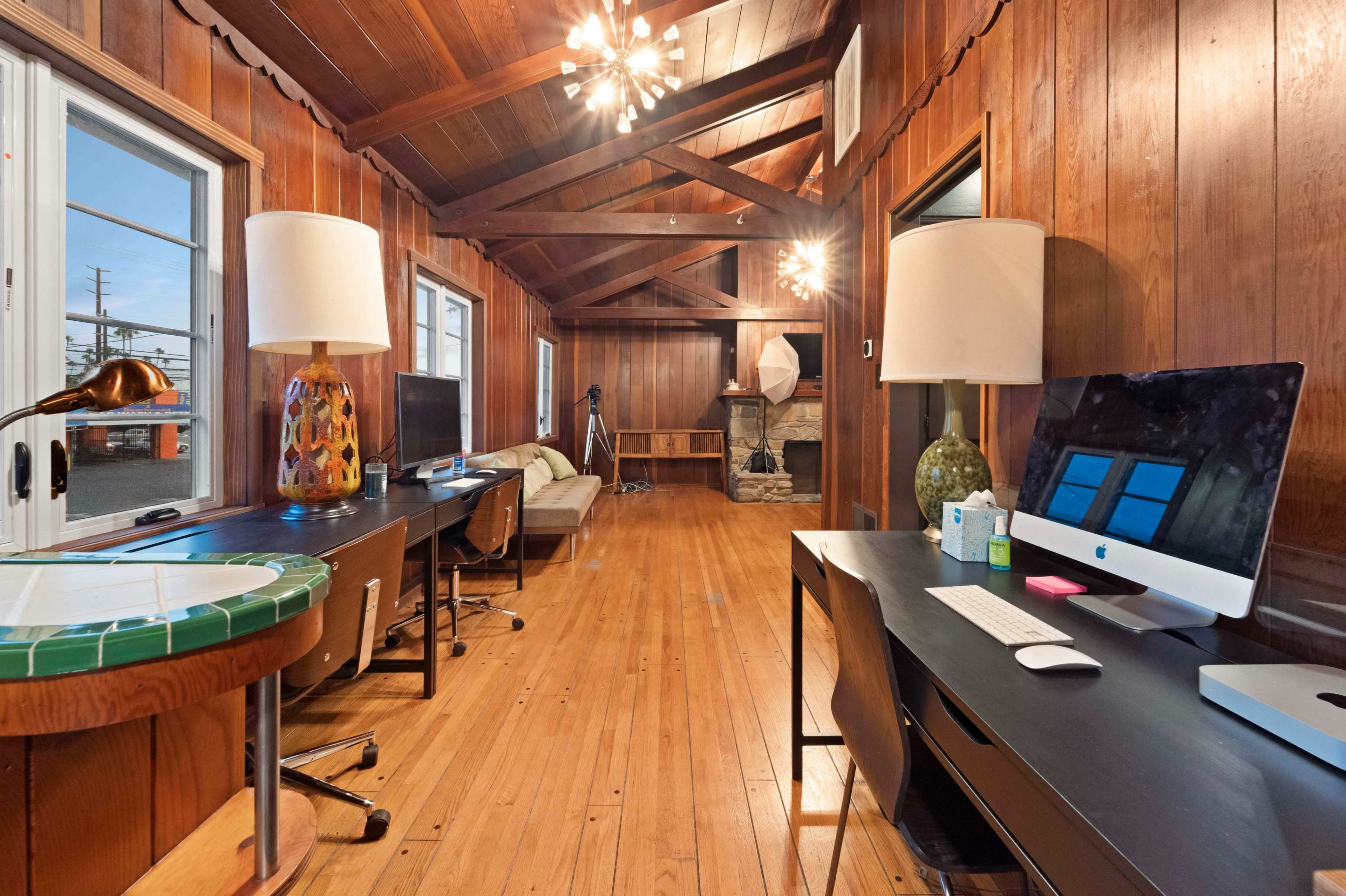 A spacious room with wooden walls and ceiling, featuring two desks with computers, a sofa, and a decorative lamp.