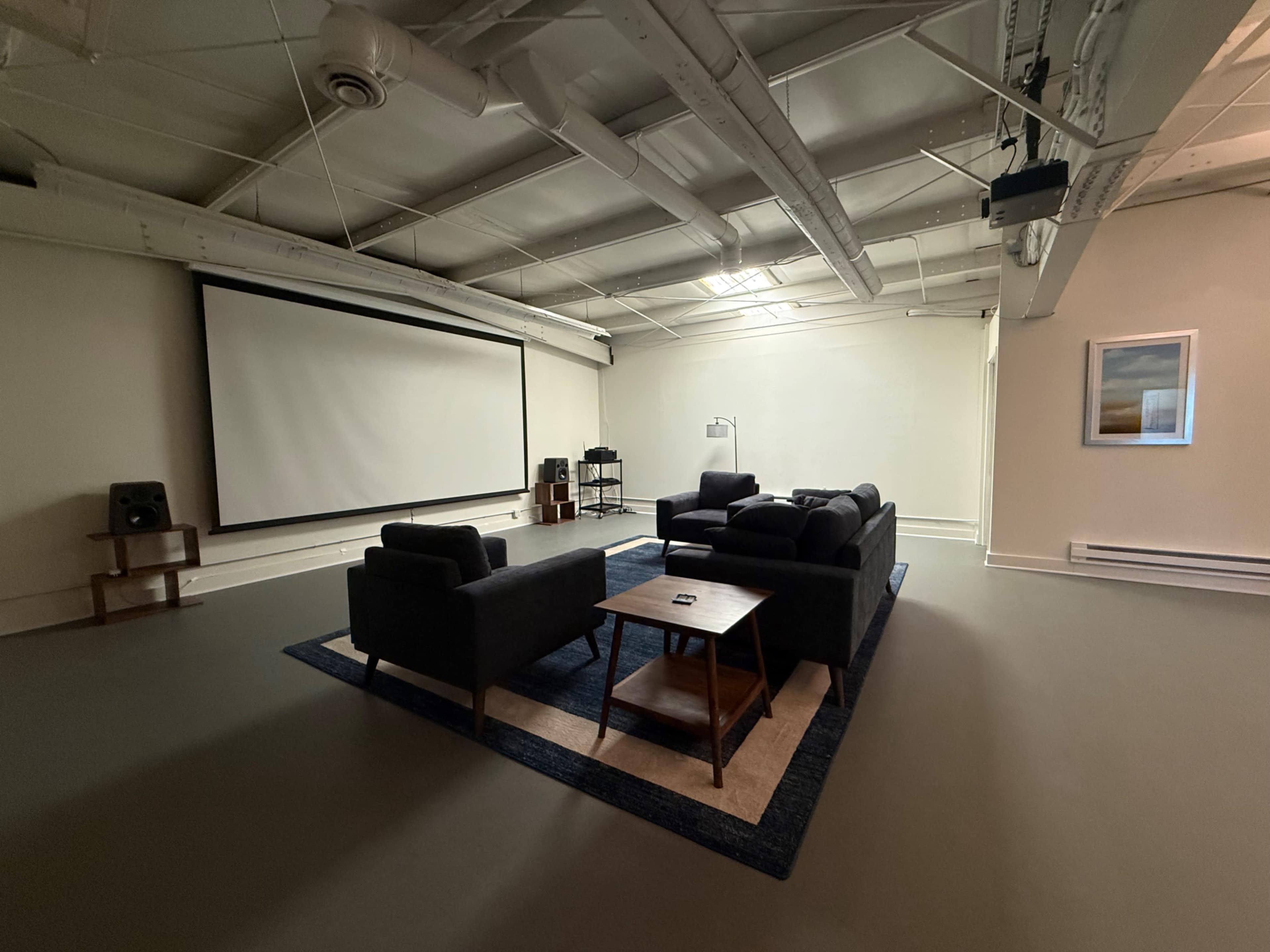 The image shows a spacious, modern room with a large screen, black couches arranged around a coffee table, and minimalistic decor.
