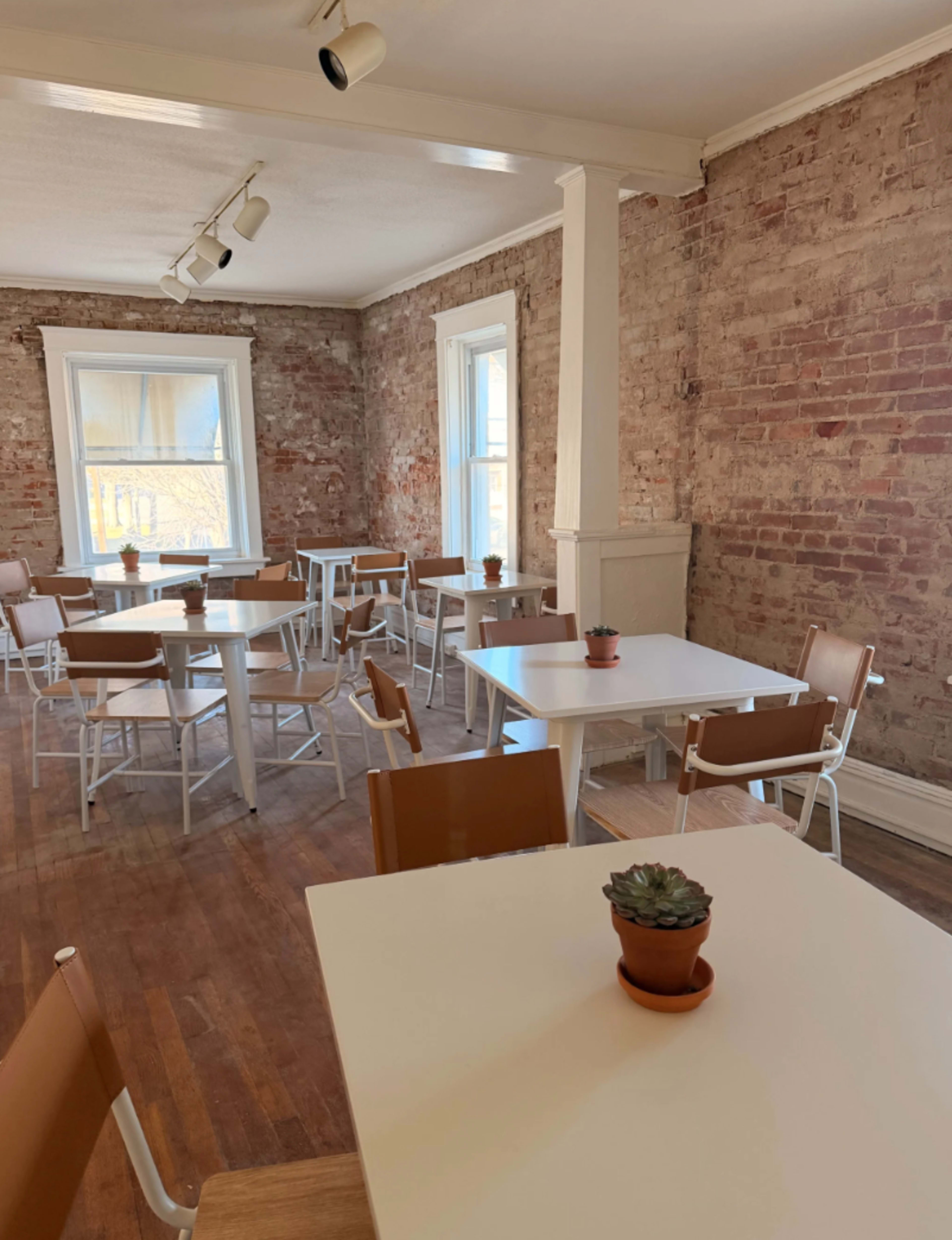 The image shows a spacious dining area with several white tables and brown chairs arranged on a wooden floor, set against a backdrop of exposed brick walls and large windows.
